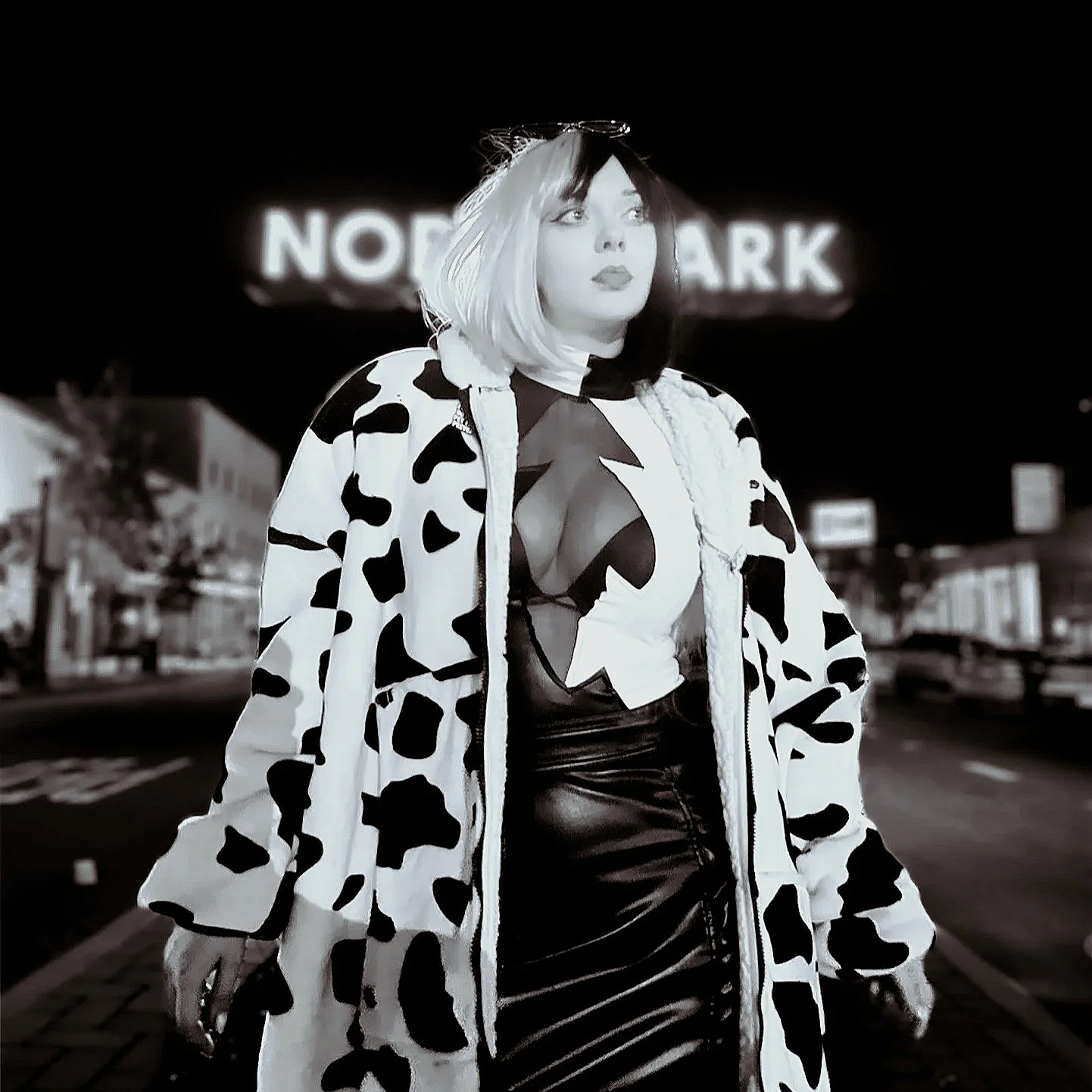 Woman with platinum blonde hair and light skin standing on a city street at night, wearing a cow print coat over a mesh top and leather skirt, with a illuminated sign reading 'NORTH PARK' in the background.