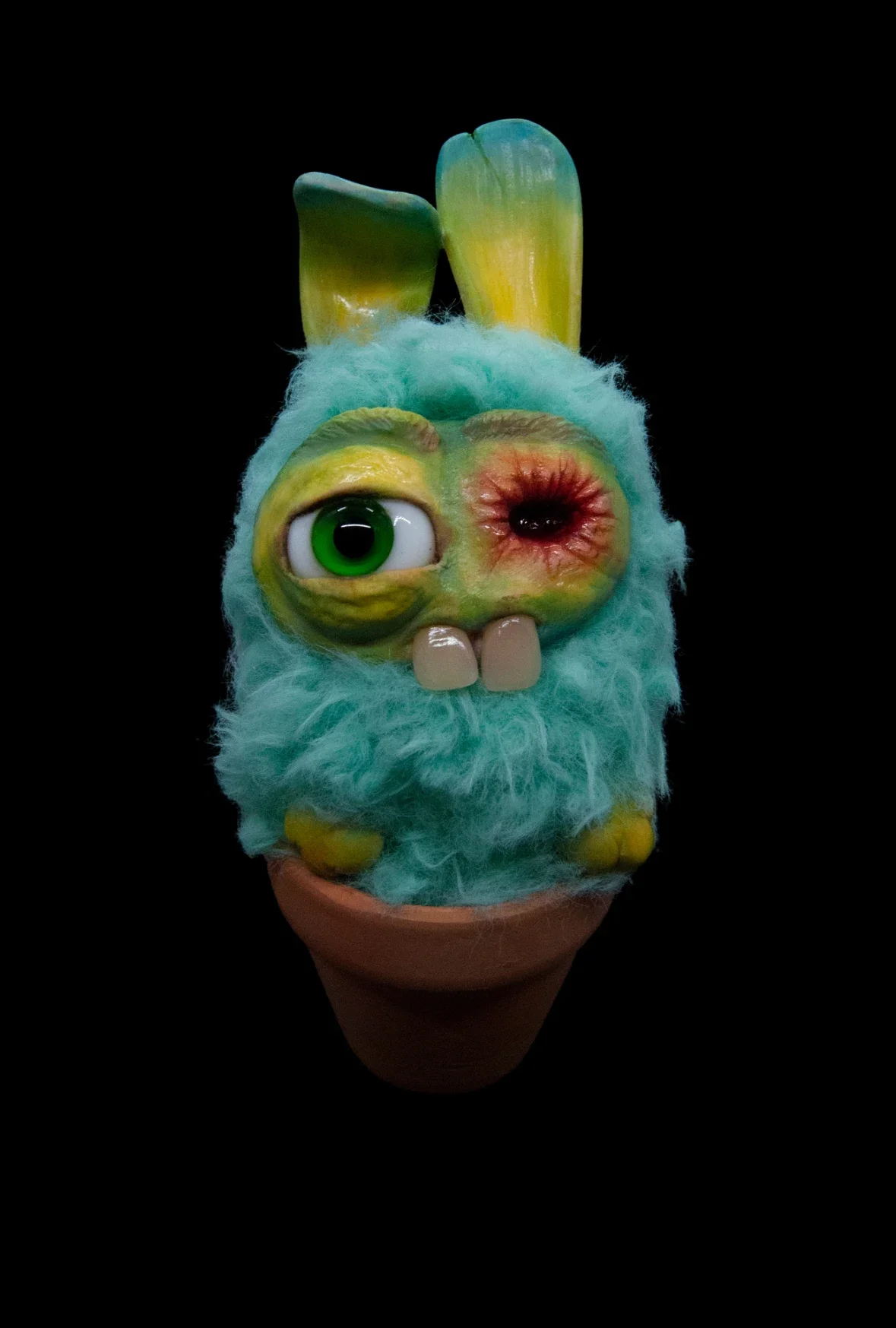A colorful, creature-like stuffed toy with one large green eye, one closed eye, large front teeth, fluffy turquoise fur, and two tall ears, sitting in a small terracotta pot against a black background.