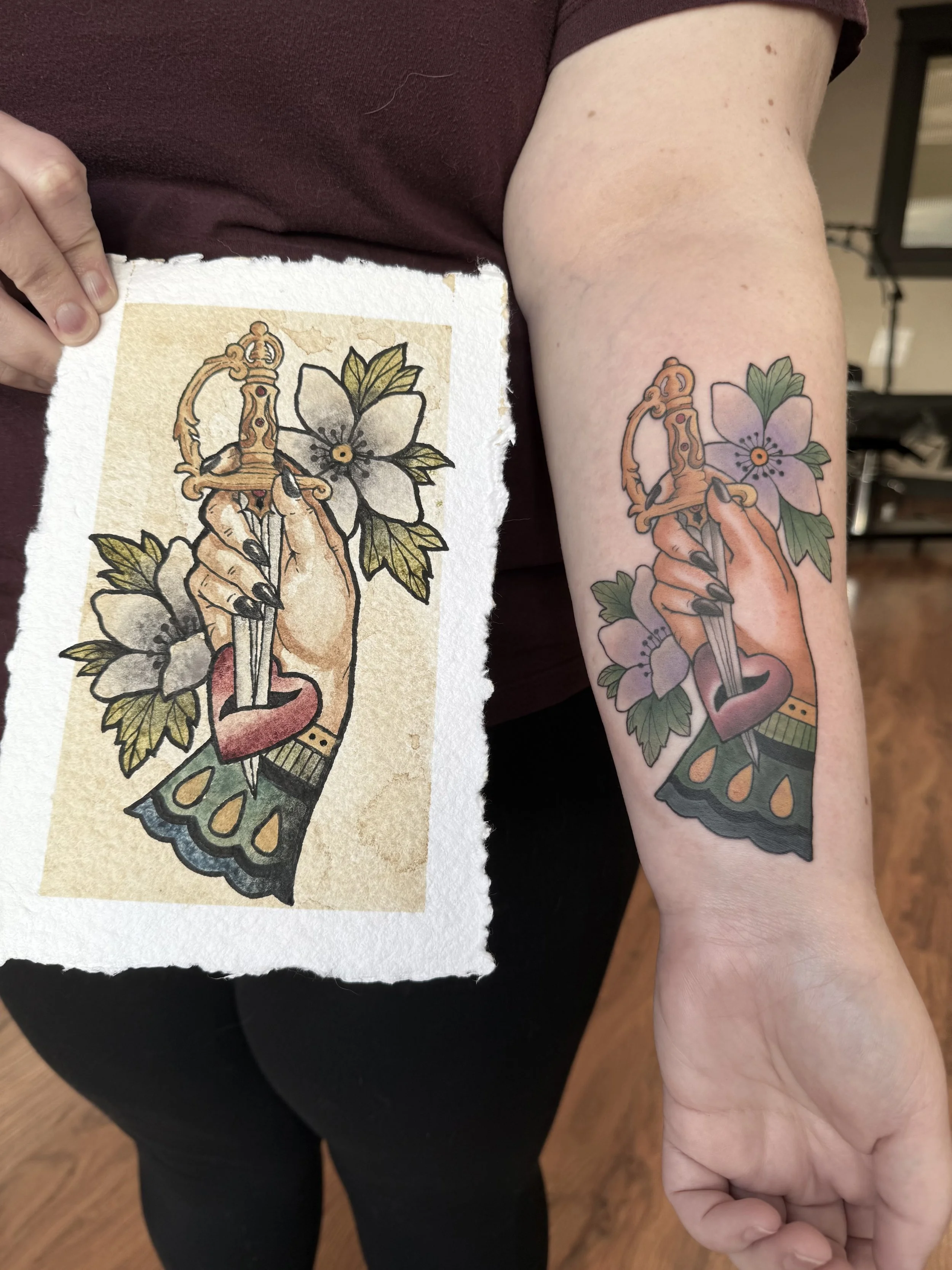 Original watercolor painting made with handmade watercolor paints beside its matching forearm tattoo, depicting a hand holding an ornate dagger surrounded by purple Oregon anemone flowers and green leaves. it depicts wearing your heart on your sleeve