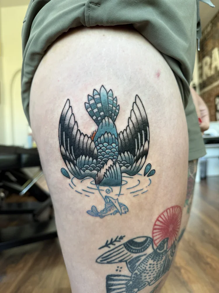 Blue and black traditional style native kingfisher bird diving into water toward a fish, tattooed on the thigh with outstretched wings. The head and fish are not colored in, only lined in blue ink.