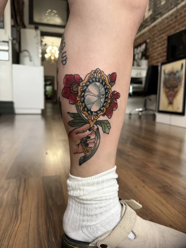 Neo-traditional calf tattoo of a hand holding an ornate gold mirror, framed by deep red hellebore flowers and green leaves.