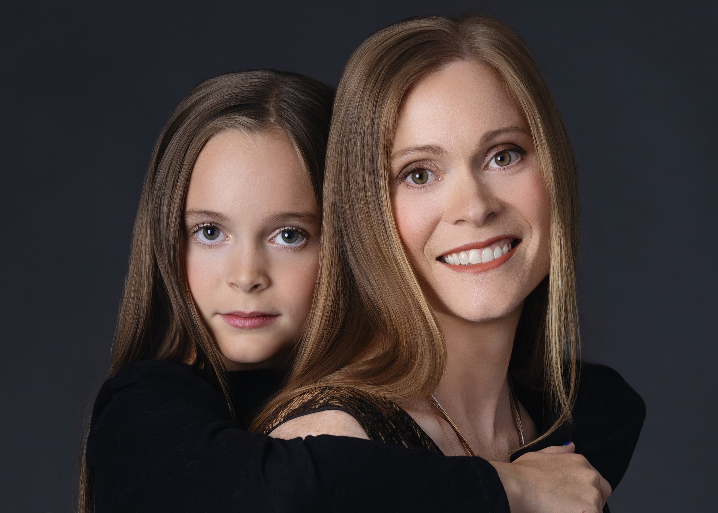 A woman with long blonde hair smiling with her arms crossed, and a young girl with brown hair hugging her from behind against a dark background.