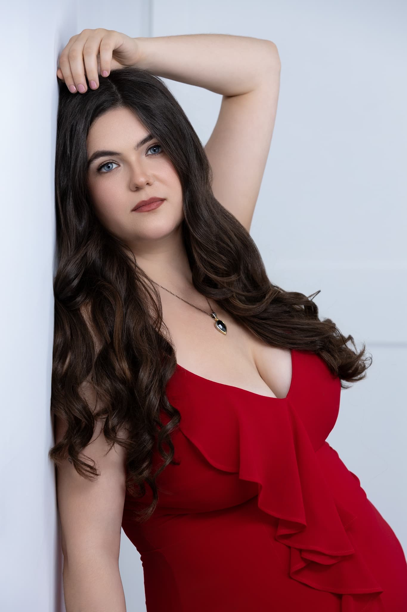 A woman with long brown hair, blue eyes, and fair skin wearing a red dress with ruffles, posing against a light-colored wall.