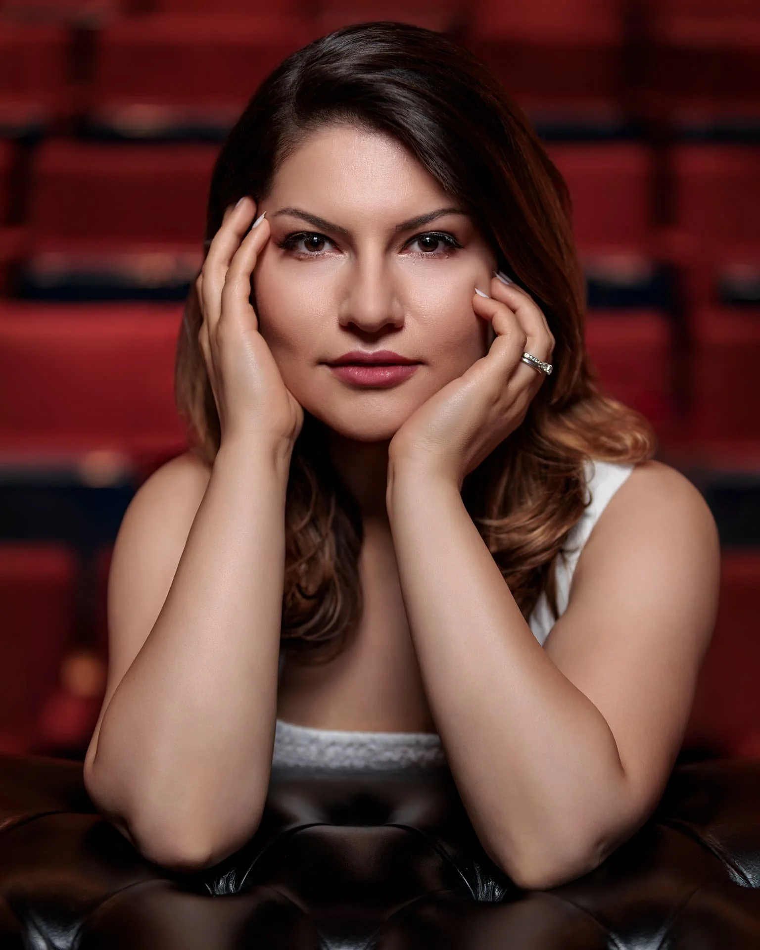 A woman with brown hair poses with her hands framing her face in a studio with a blurred red and black background.