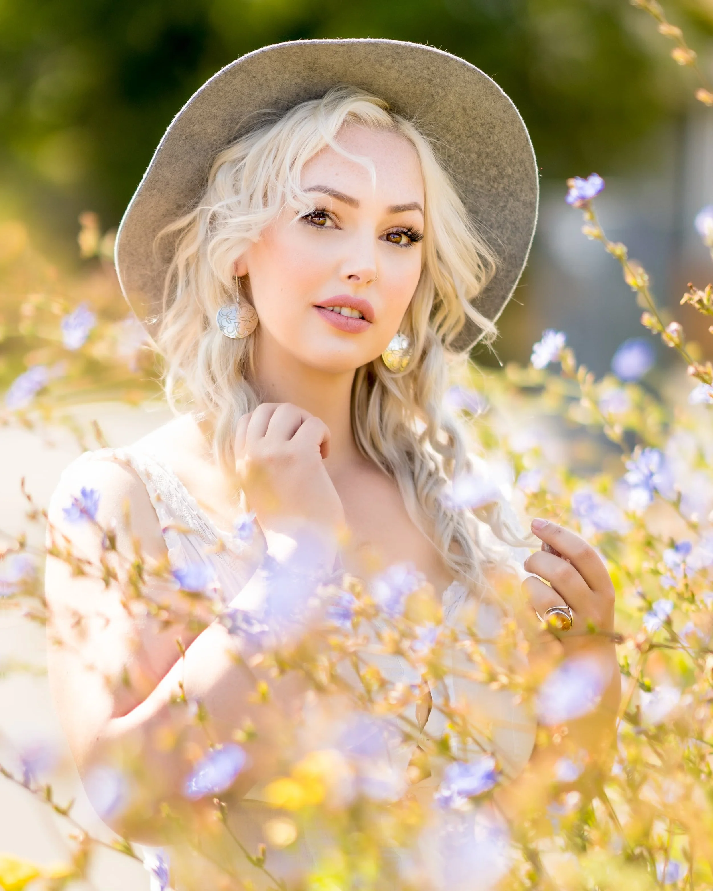 A woman with blonde curly hair, wearing a wide-brimmed hat and patterned earrings, standing amidst purple flowers outdoors.