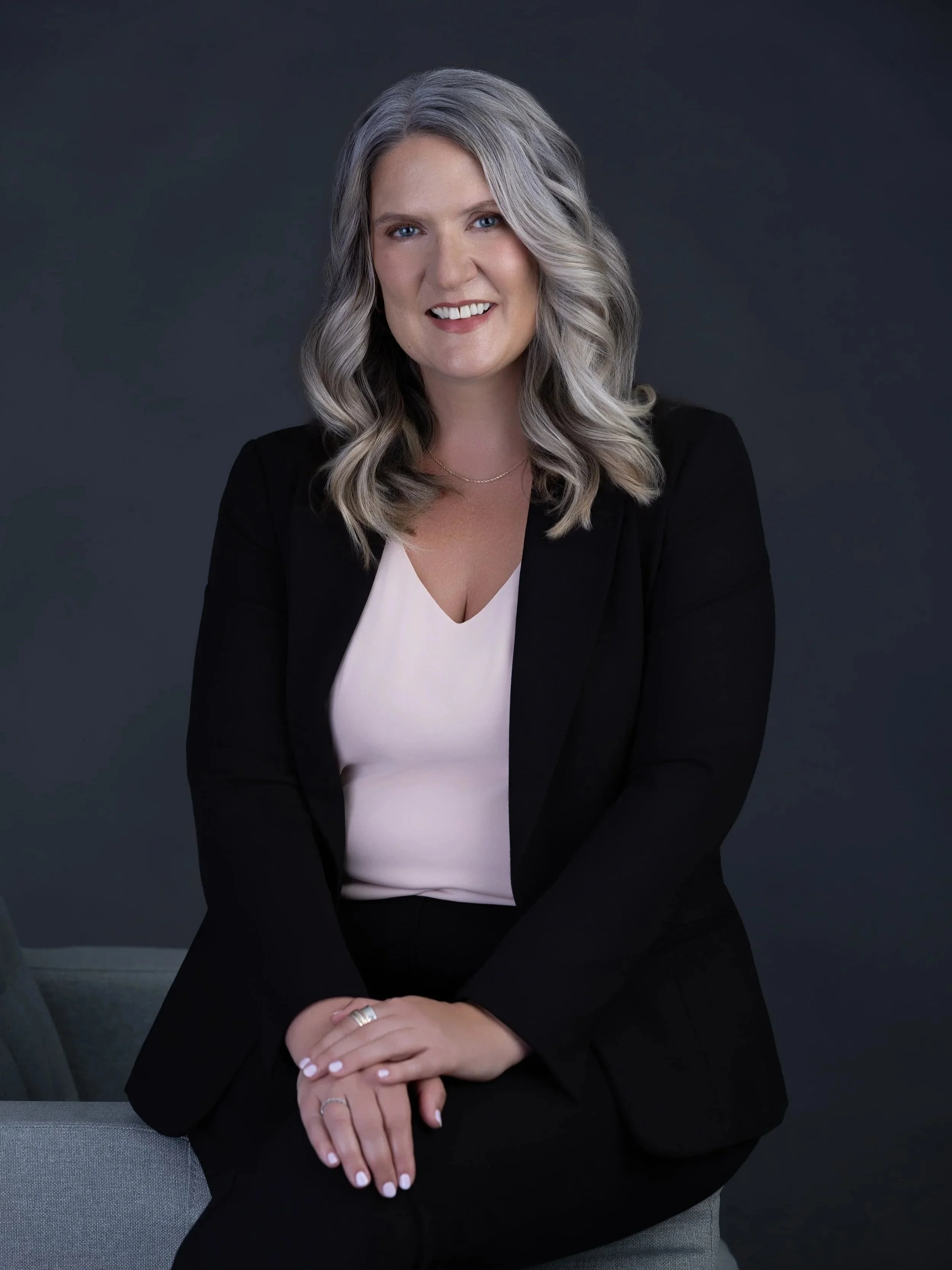 Glenn Mathieson Best photographers in Duncan Mill Bay CowichanA woman with shoulder-length gray hair and blue eyes, smiling, wearing a black blazer over a white top, sitting on a gray chair against a dark background.