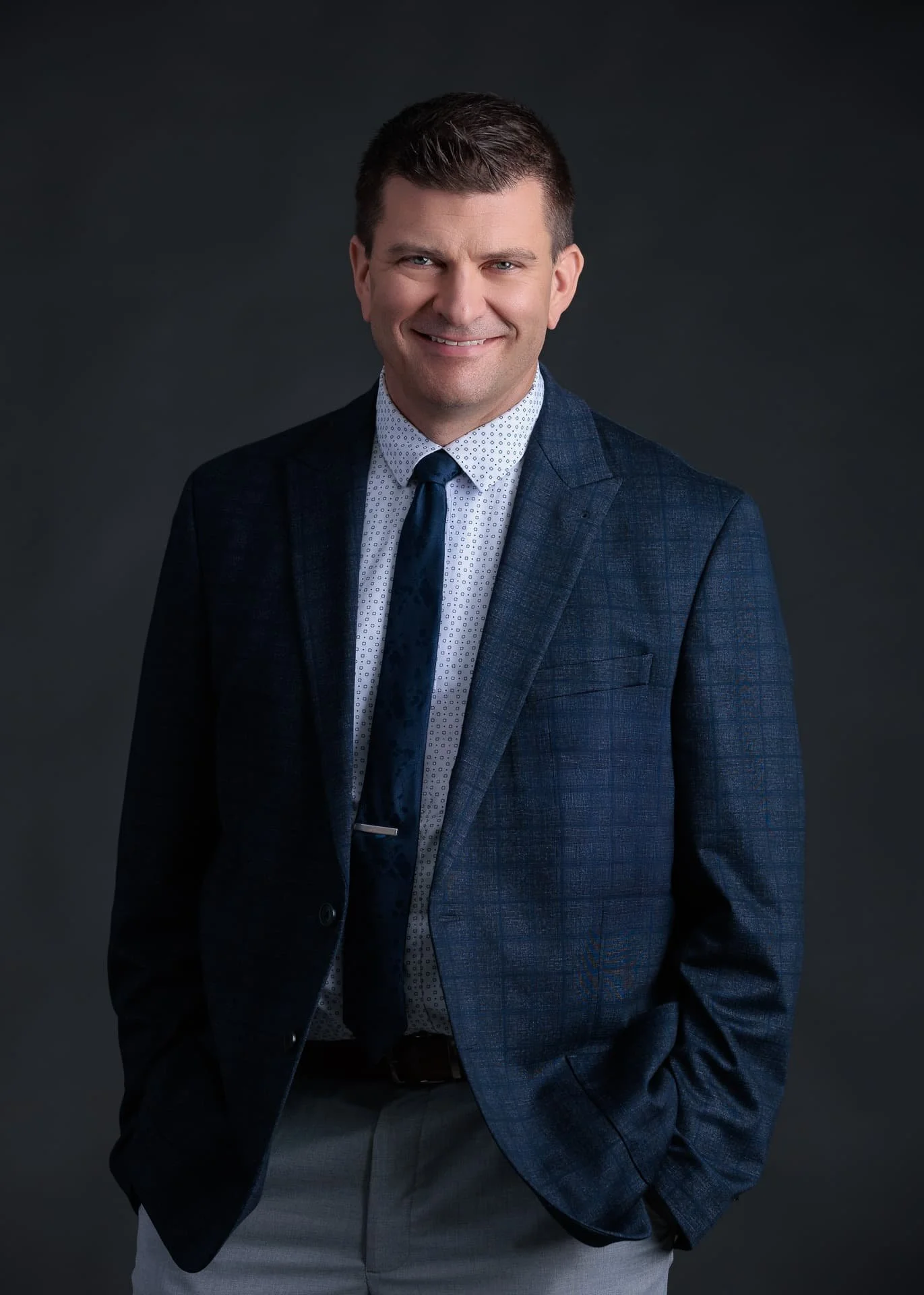 Headshot Portrait of a smiling man in a dark blue suit, white dress shirt with small pattern, and dark tie, posing against a dark background. Glenn Mathieson