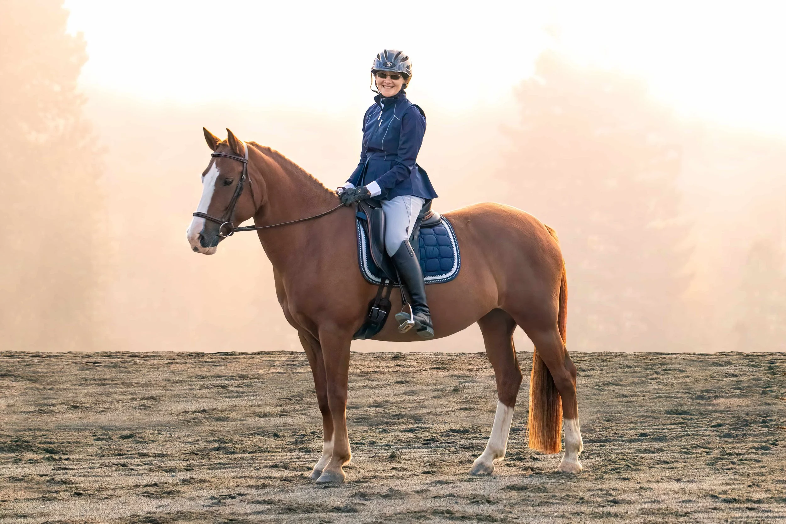 A smiling woman in riding gear, including a helmet and gloves, sitting on a brown horse with white markings on its face and legs. The horse is standing on a flat, sandy surface at sunset or sunrise.