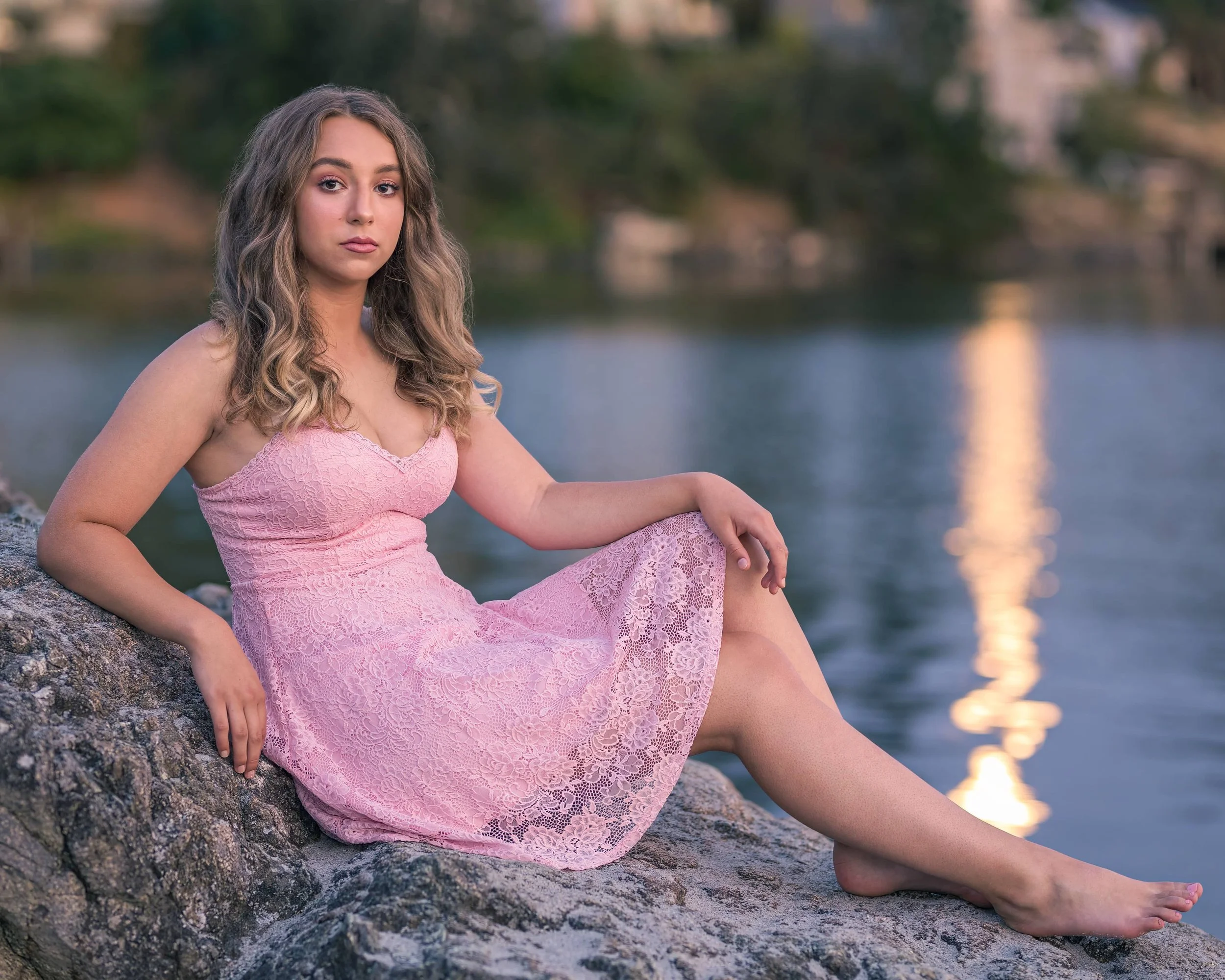 A young woman in a pink lace dress sitting on a rock by a lake at sunset, looking at the camera.