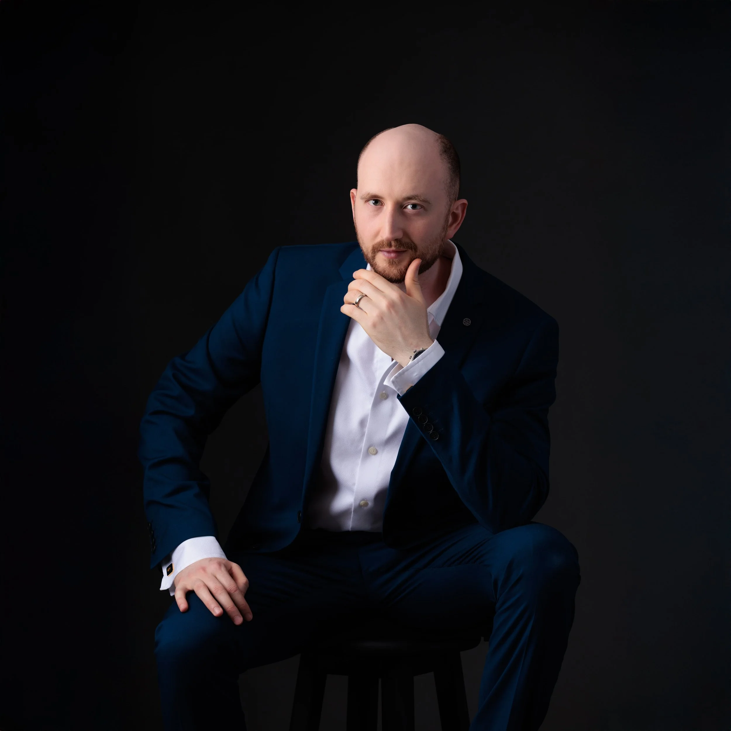 A man in a dark blue suit sitting on a stool against a black background, with his hand on his chin, looking thoughtfully at the camera.