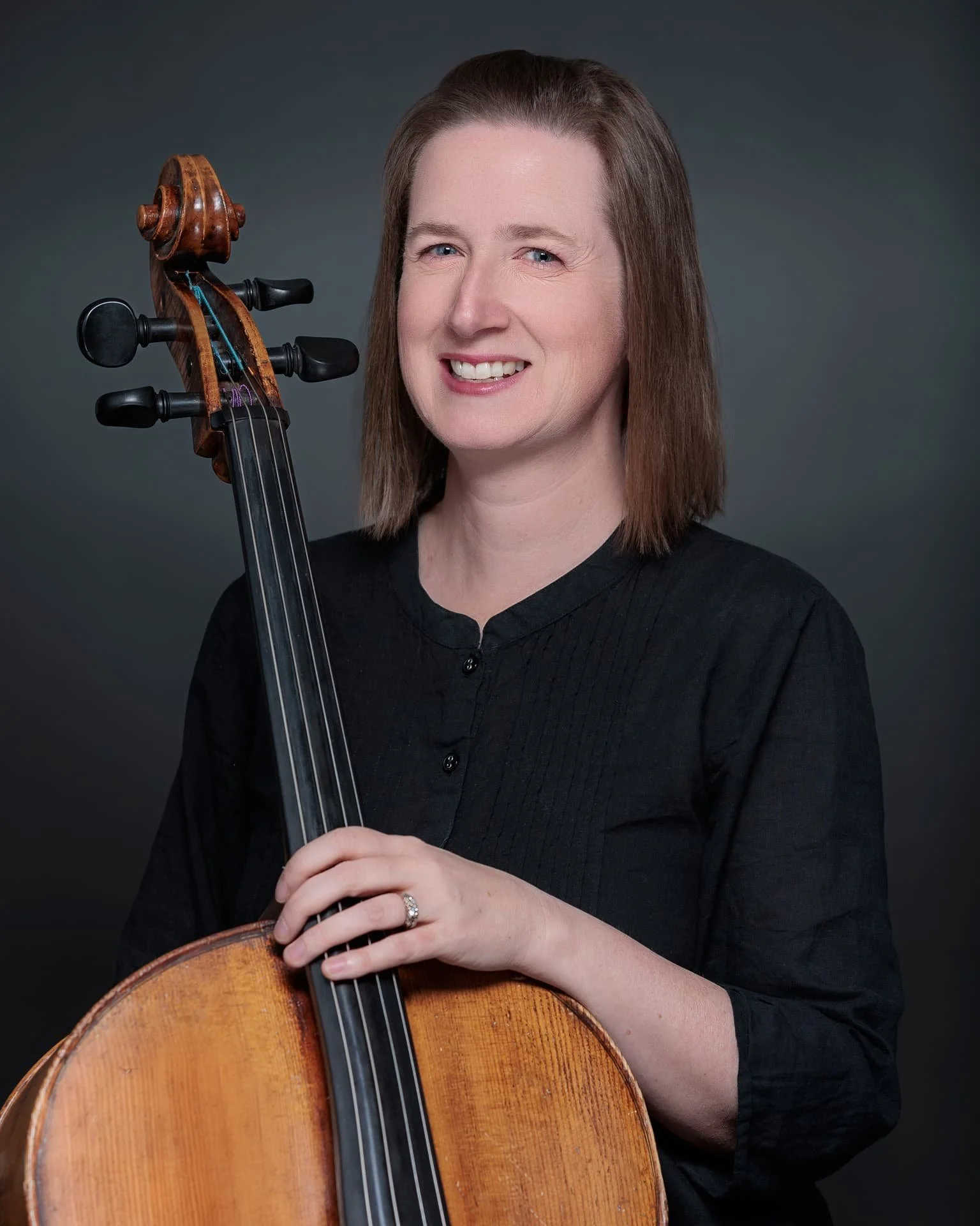 A woman with shoulder-length brown hair holding a cello and smiling at the camera against a dark gradient background.