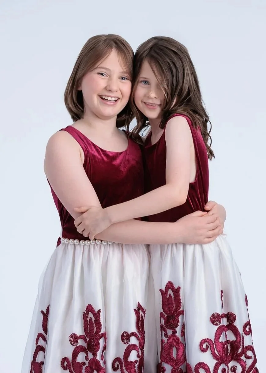 Two young girls wearing matching red velvet tops and white skirts with red embroidery, hugging each other and smiling.