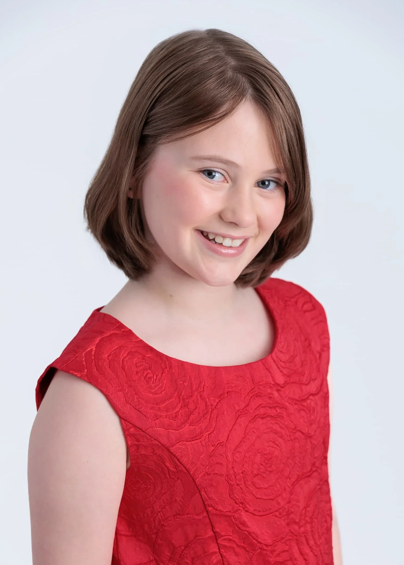 A young girl with long brown hair sitting on a white platform, wearing a black faux fur shawl, a patterned black and white skirt with floral designs, and black shoes, against a plain white background.