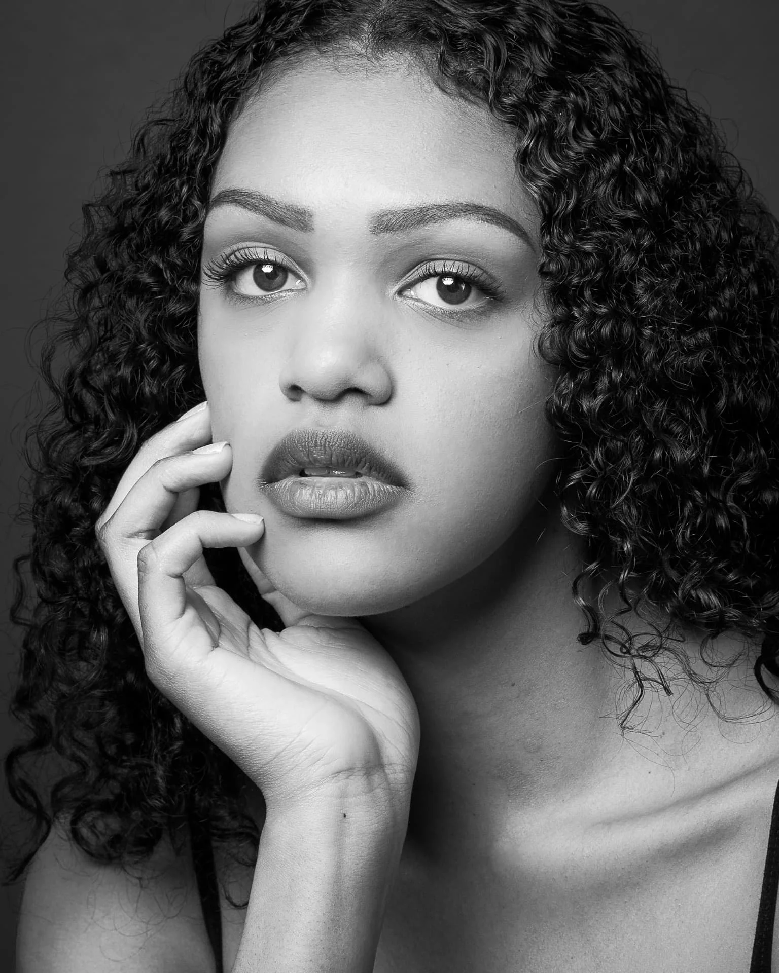 Black and white portrait of a woman with curly hair, slightly touching her face with her hand, looking directly at the camera.