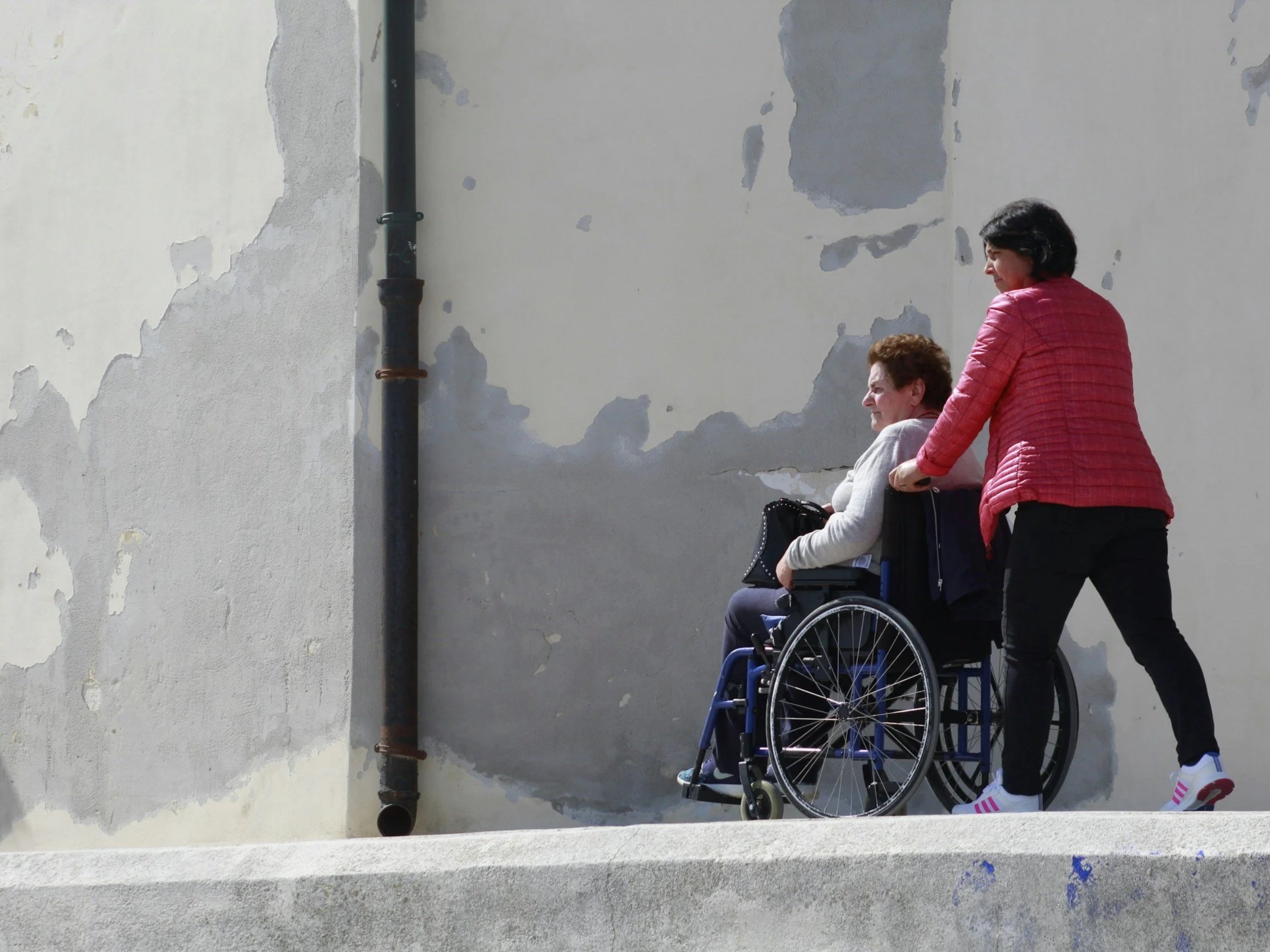 A woman in a wheelchair being pushed by another woman along a sidewalk in front of a weathered wall.