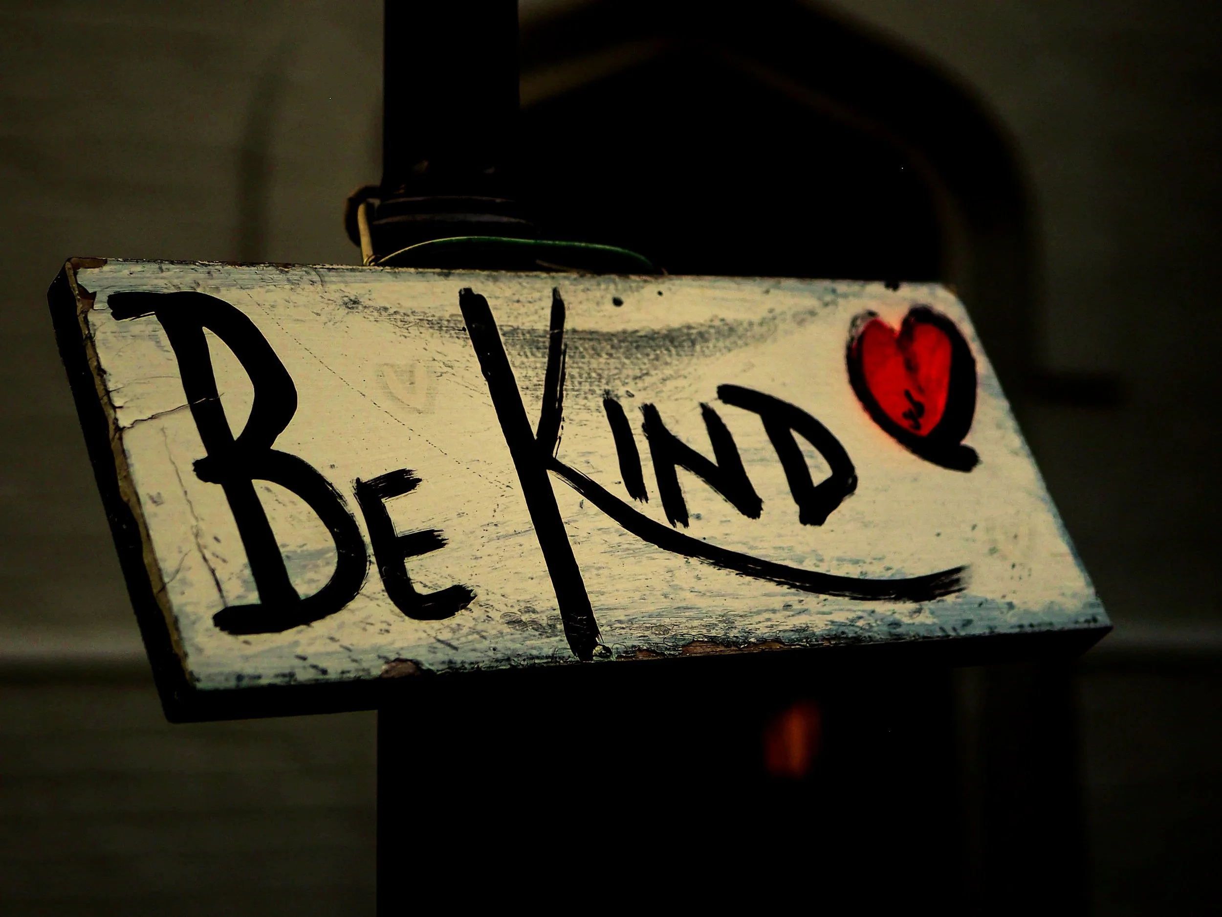 Wooden sign with the words "BE KIND" in black letters, a small red heart drawn above the letter "D".