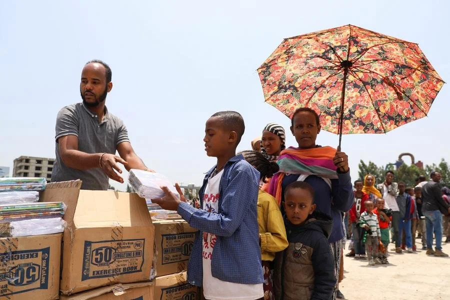 People stand in line to receive supplies from a man at an outdoor distribution event, with a woman holding a floral umbrella and children waiting nearby.