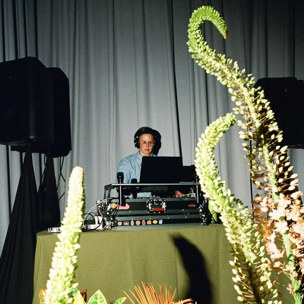 A woman DJ wearing headphones, standing behind a DJ setup with a laptop, in front of a stage with a gray curtain backdrop and large black speakers, with decorative green and pink flowers in the foreground.