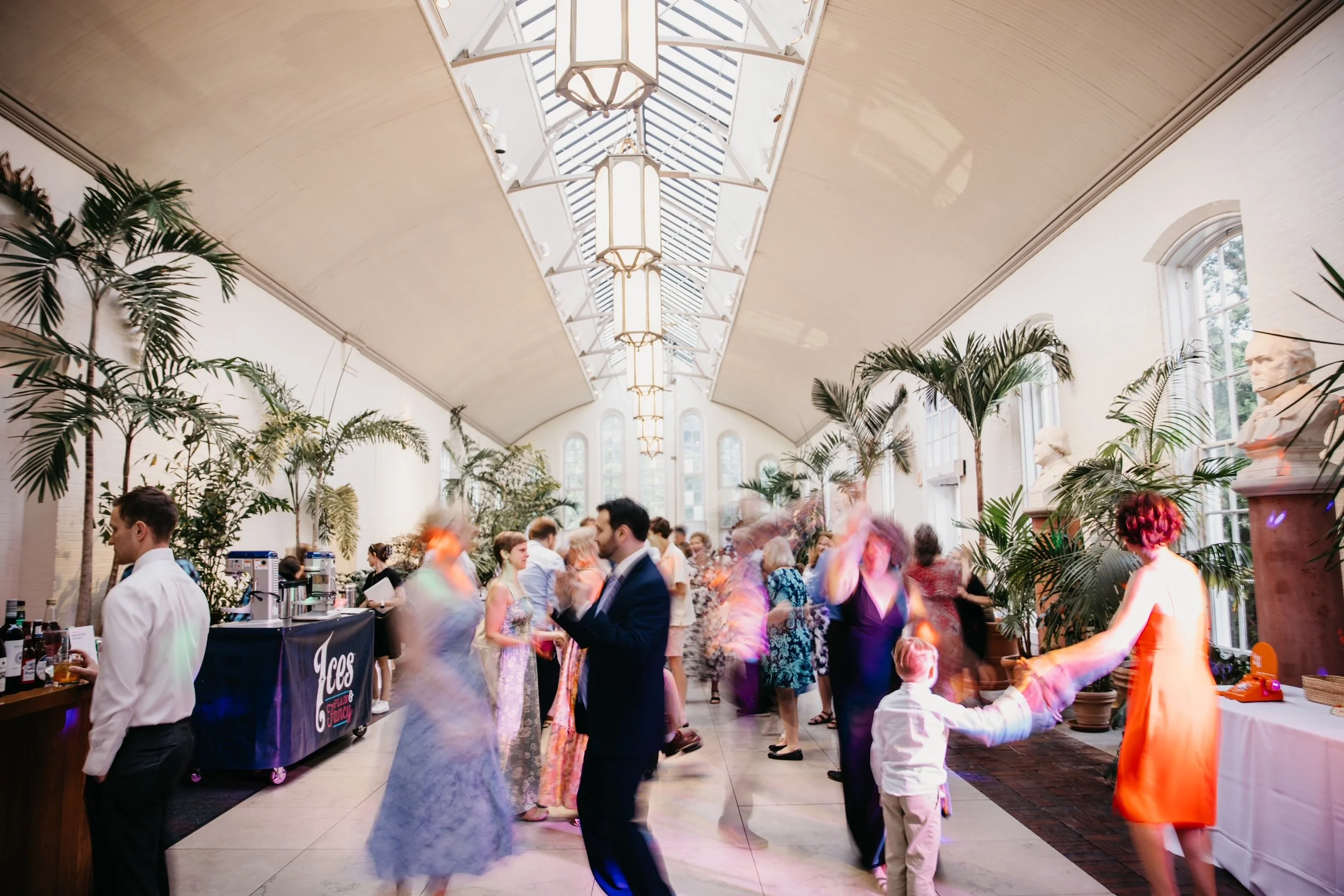 People dancing and socializing in a bright, spacious indoor venue with tall windows, lush green plants, and elegant chandeliers, during a festive event.