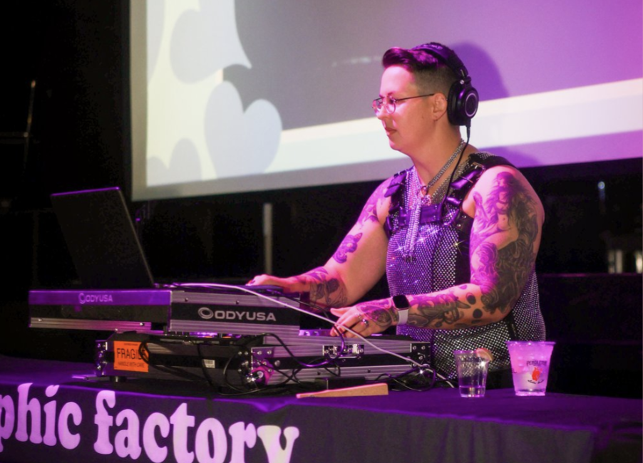 A woman with tattoos wearing glasses and headphones DJing at a music event, with a mixing console in front of her, a laptop, and drinks on the table, with a colorful screen background.