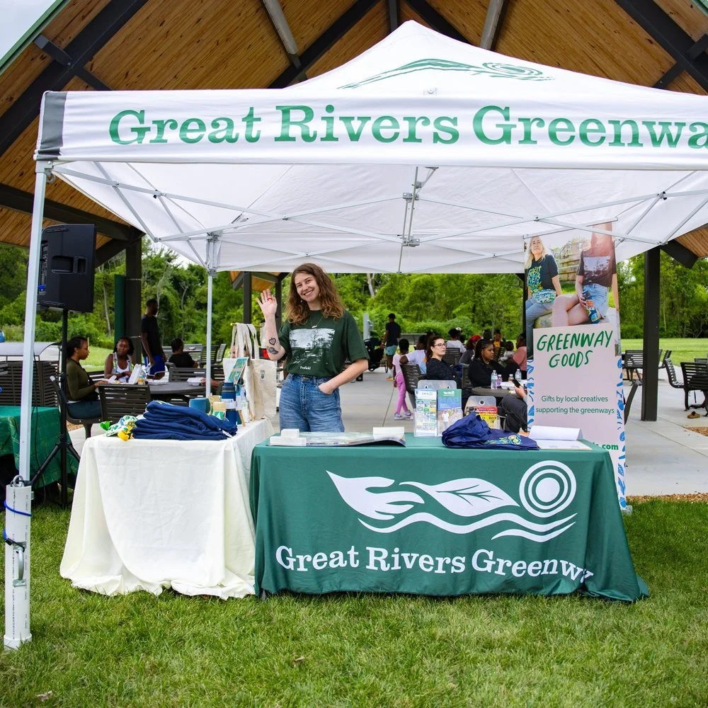A woman stands under a white canopy tent with green text reading "Great Rivers Greenway." She is smiling and waving at a community event, with a table displaying merchandise and informational materials. In the background, there are people sitting on benches and trees in a park setting.