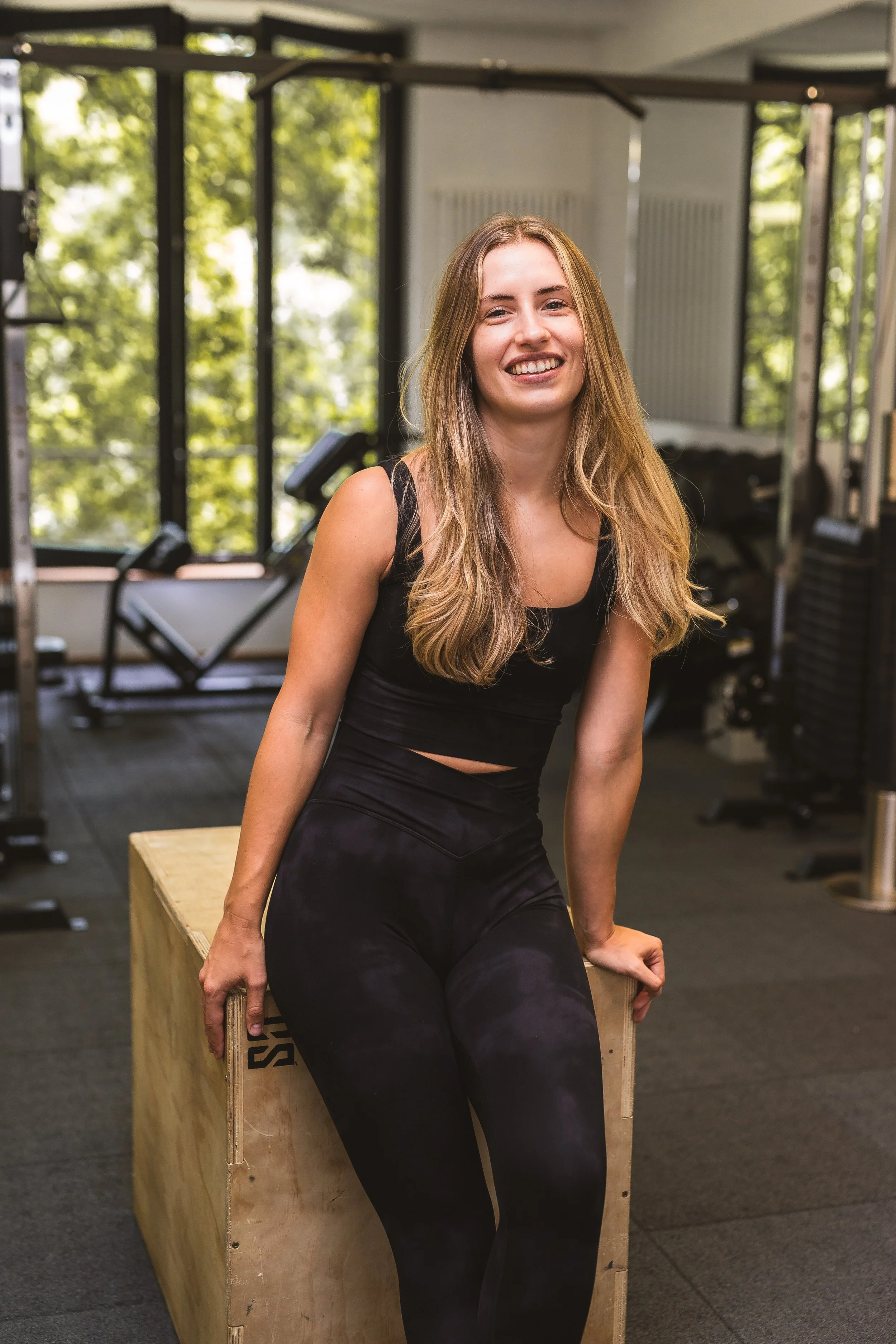 Eine junge Frau in Sportkleidung sitzt lächelnd auf einer Holzbox in einem Fitnessstudio.
