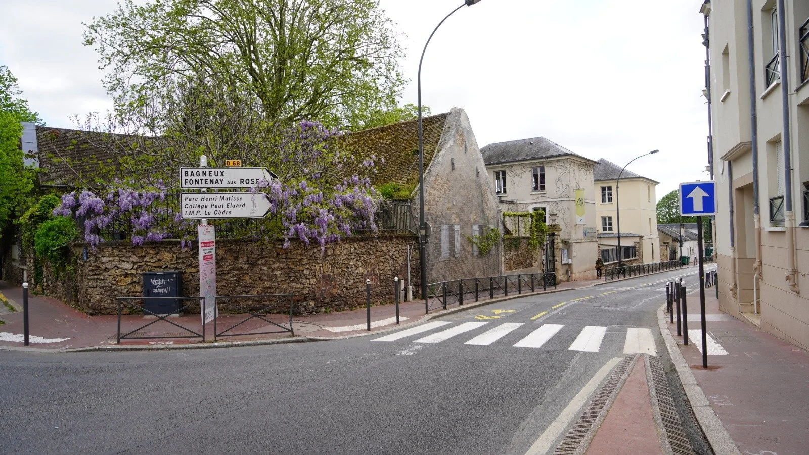 Une rue calme avec un passage pour piétons, des bâtiments résidentiels et une signalisation directionnelle, des arbres en fleur entre les bâtiments.