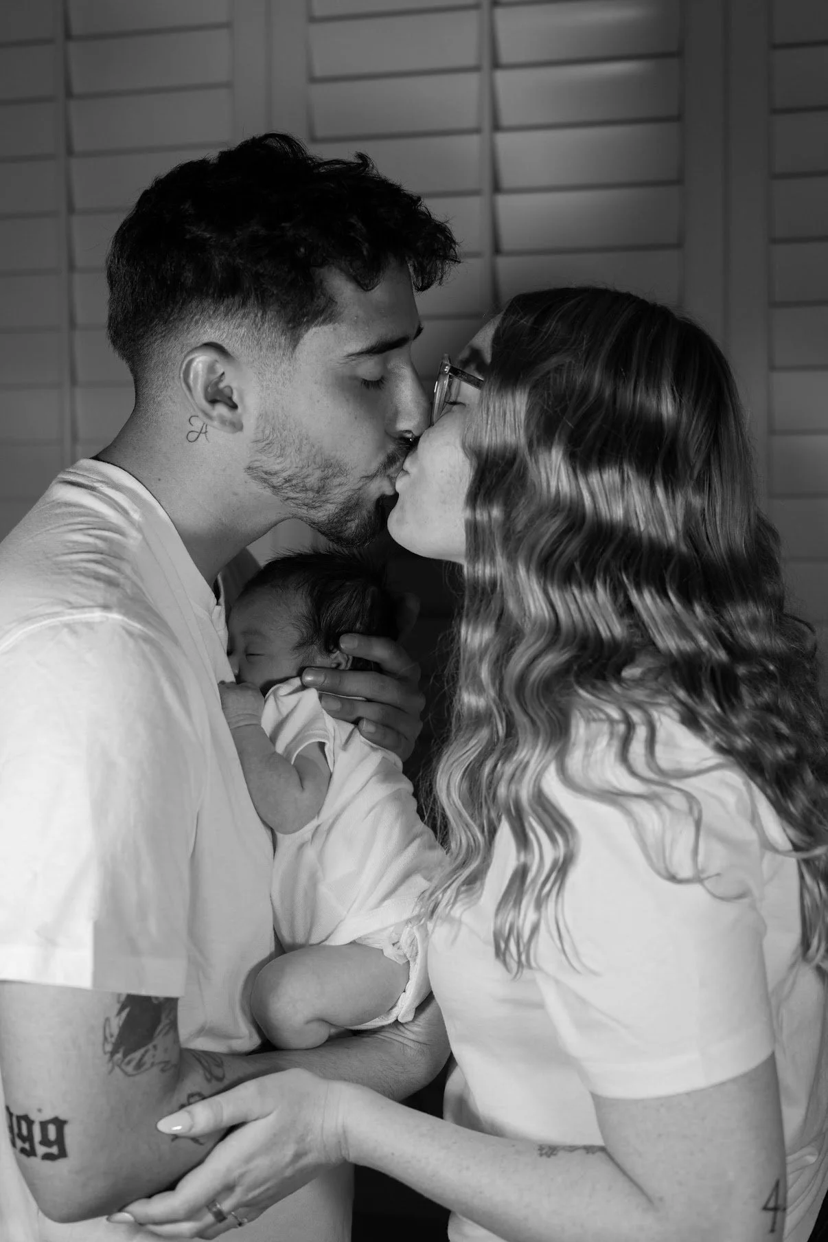 A couple kisses while holding a newborn baby between them in black and white.