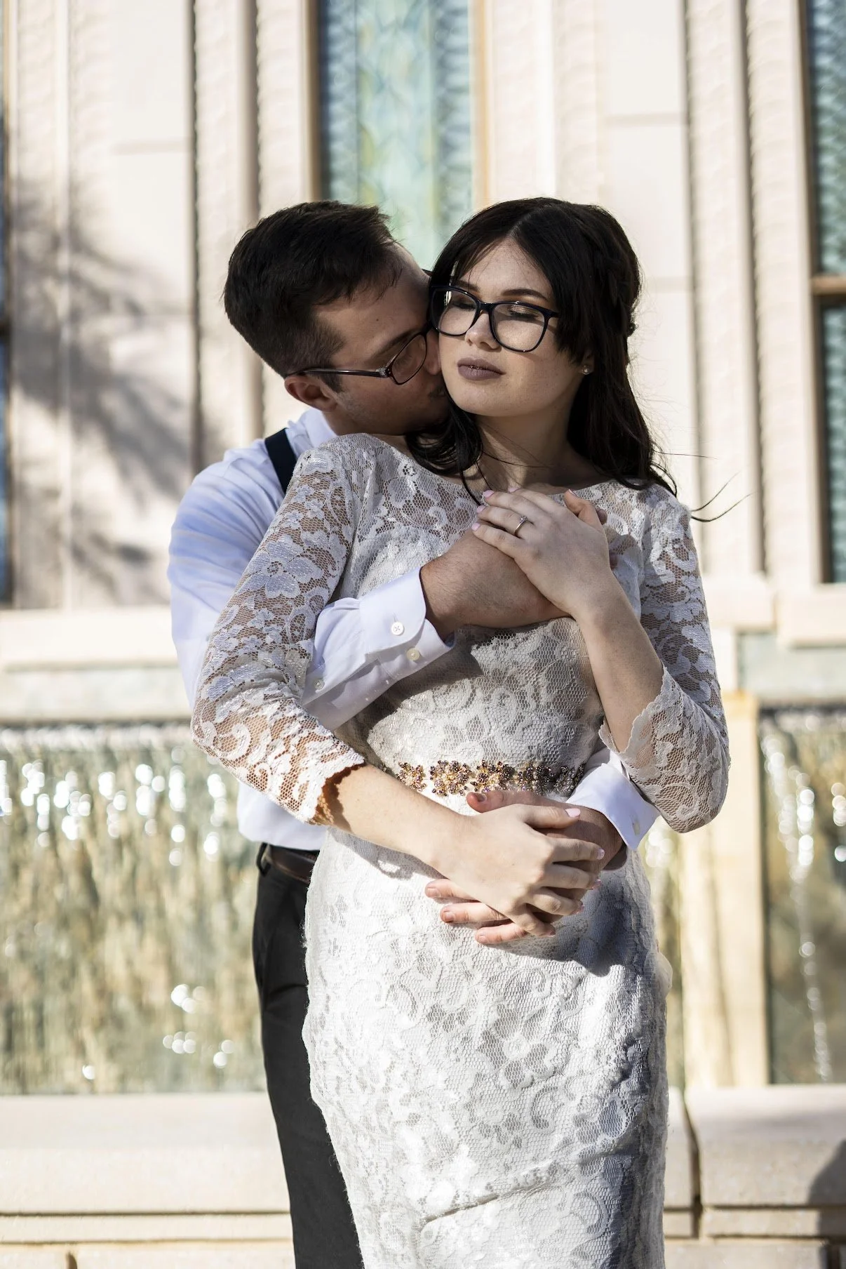 A man in white shirt and glasses embraces a woman in a white lace dress and glasses, kissing her cheek outside in front of a modern building with glass windows.