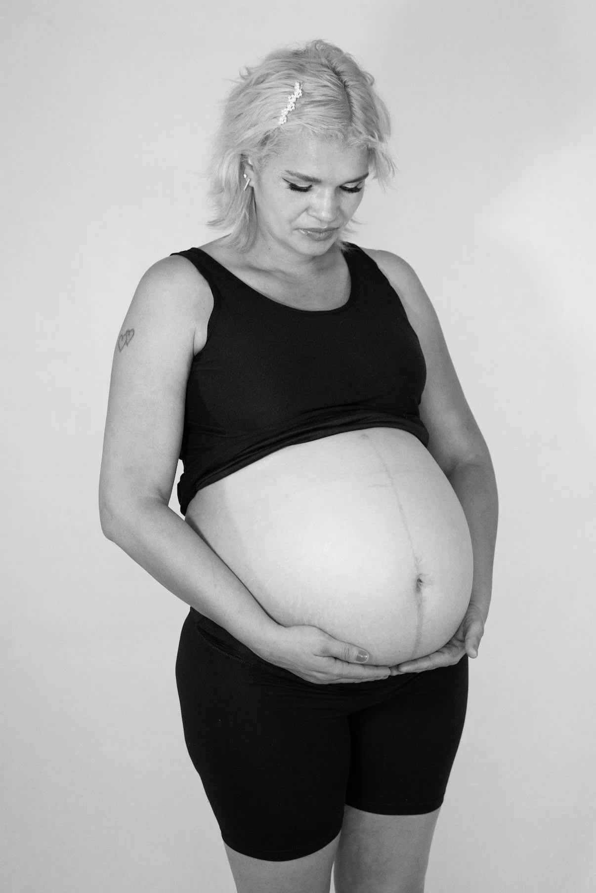 Pregnant woman with short blonde hair, wearing a black tank top and black shorts, cradling her large belly while looking down, against a plain light background.