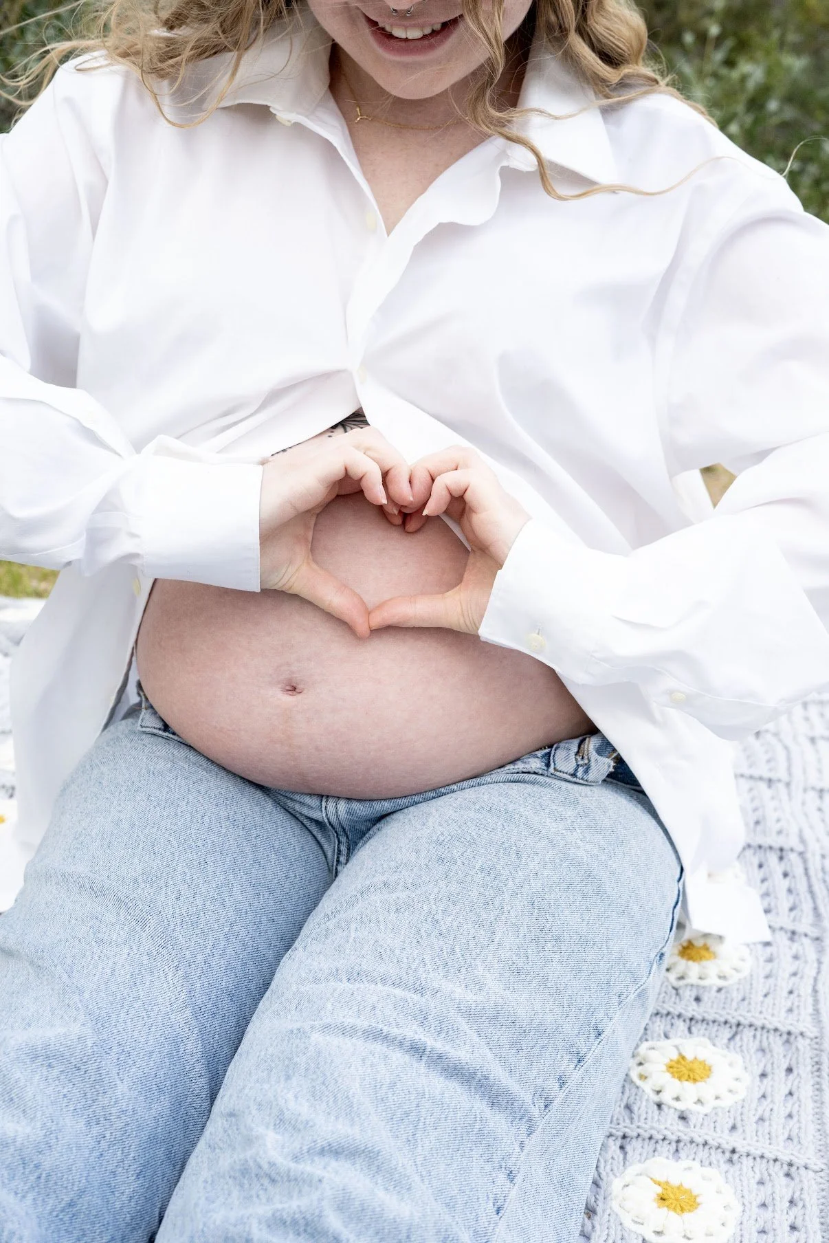 A pregnant woman wearing a white shirt and blue jeans sitting outdoors, forming a heart shape on her belly with her hands, which are also forming a heart, smiling cheerfully.