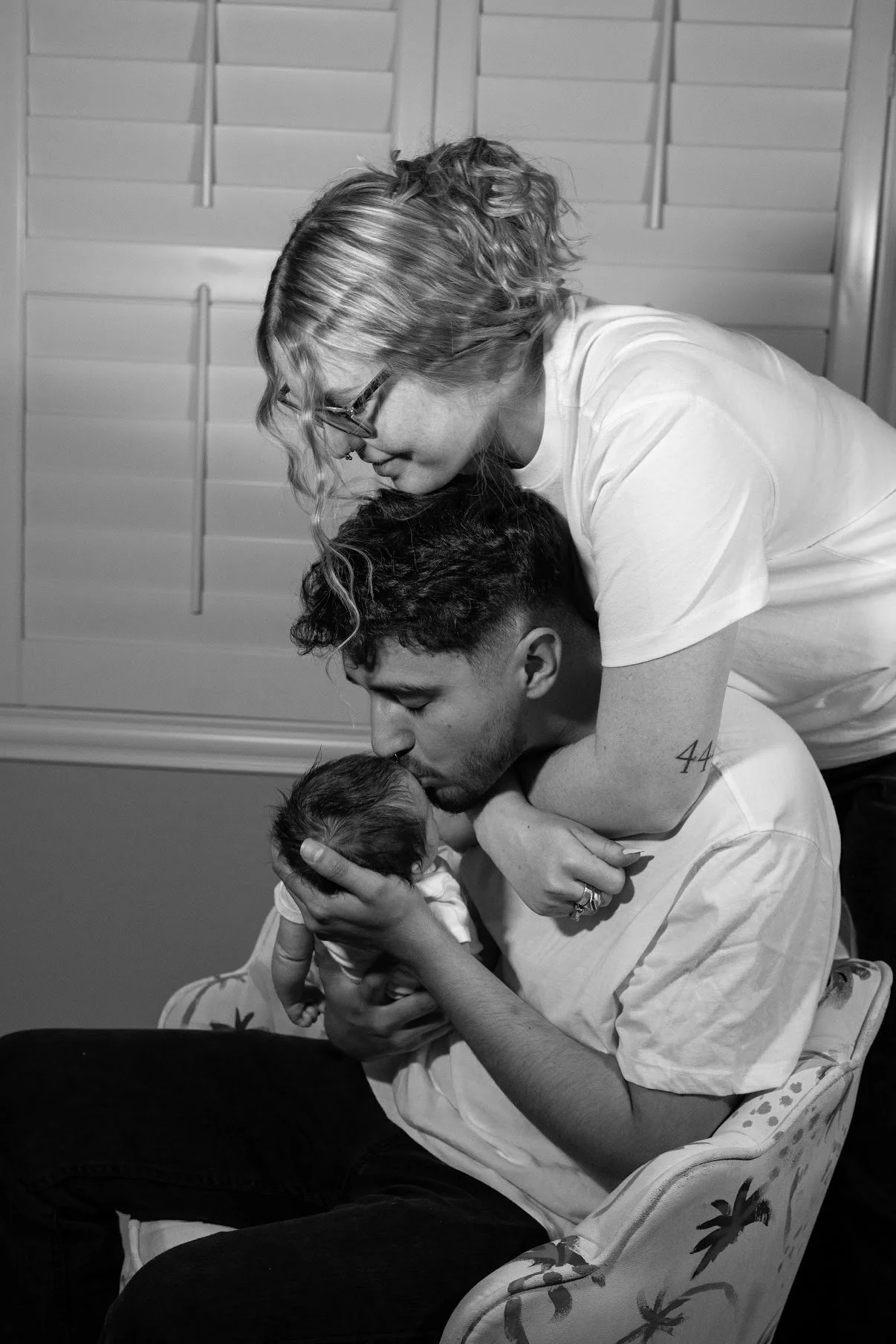 A woman and a man, both smiling and affectionate, share a tender moment holding a newborn baby. The woman leans over the man who is seated in a floral-patterned chair, gently kissing the baby's head. The scene is in a room with closed window shutters