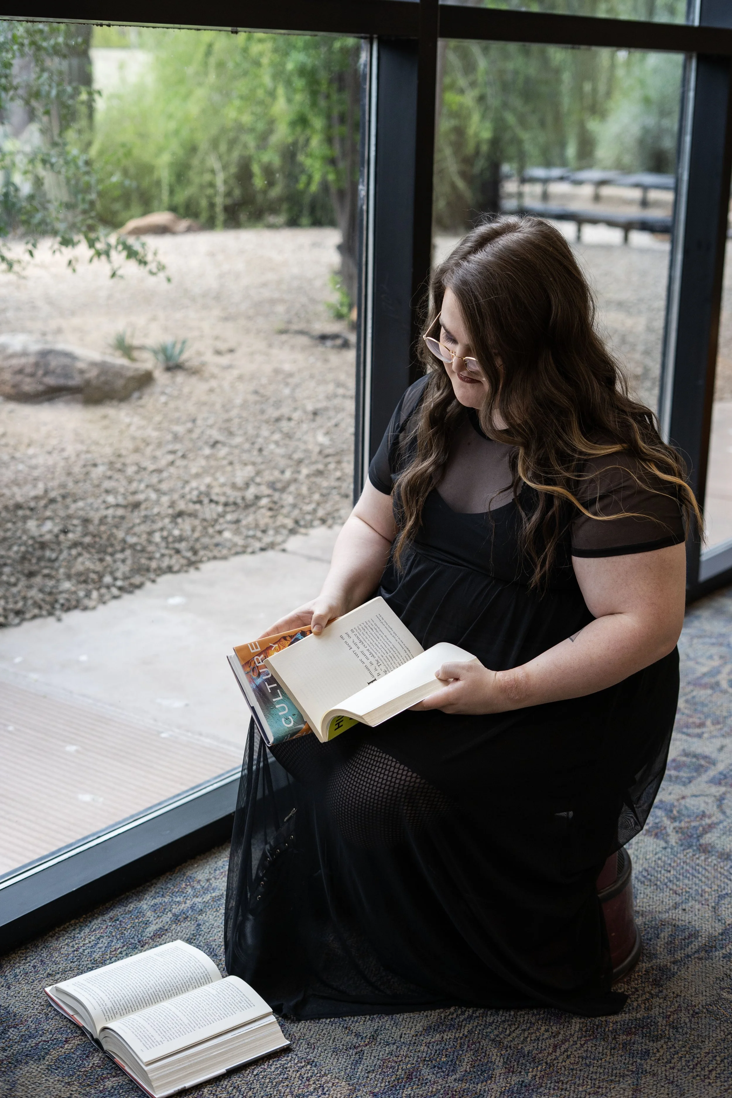A woman with long, wavy brown hair and glasses reading a book while seated on a small stool near a large window, with another open book on the carpeted floor beside her.