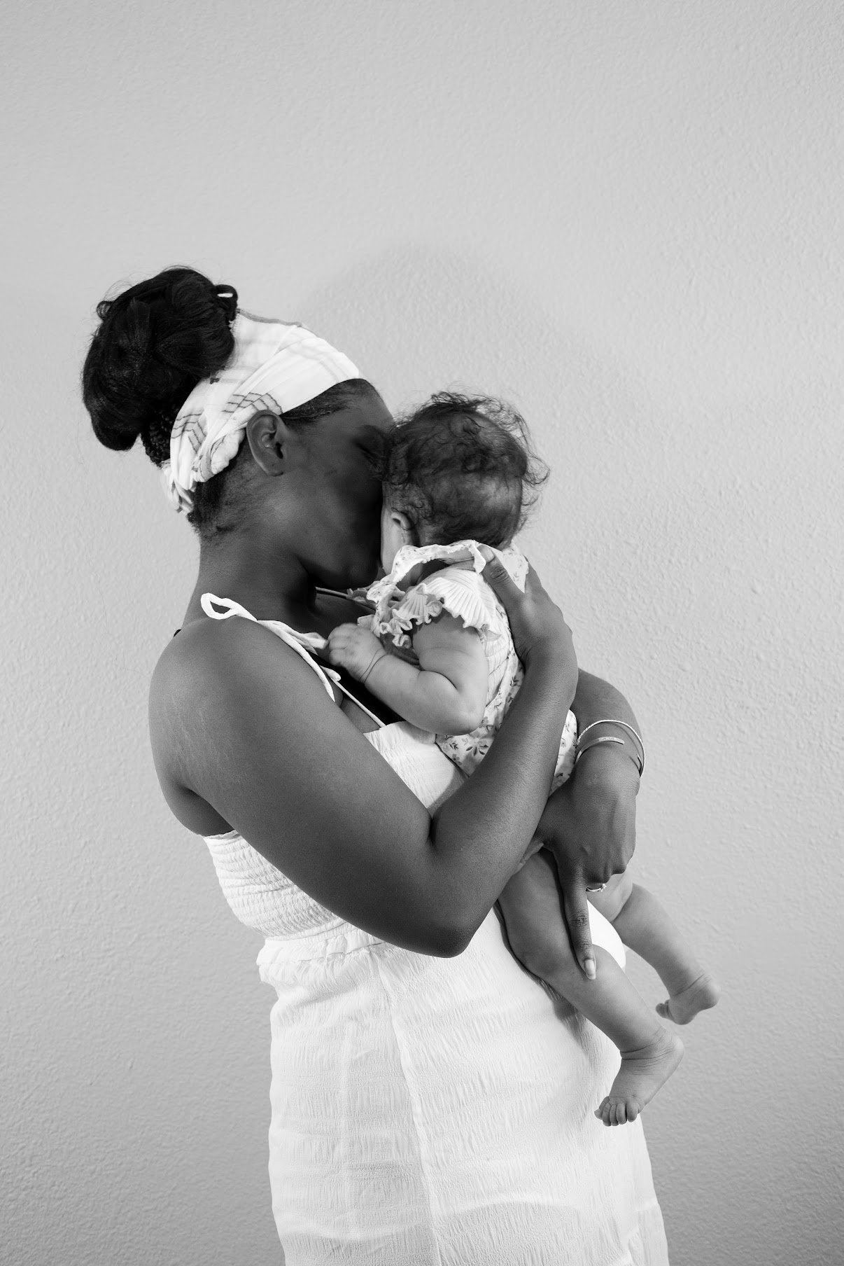 Black woman holding and kissing a young child in front of a plain wall.