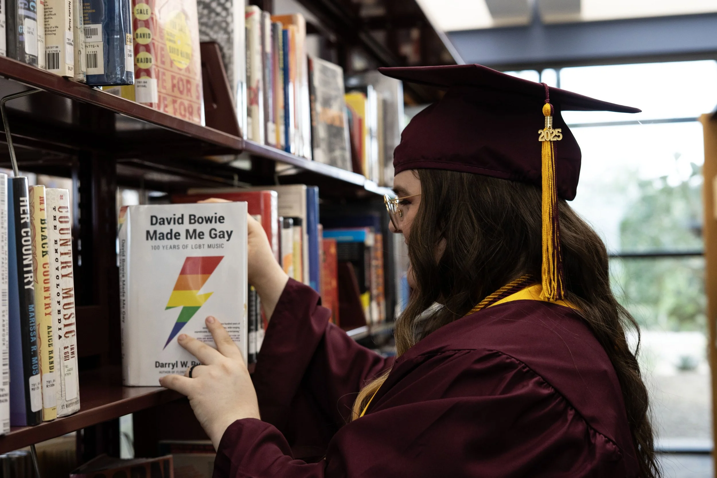 Graduate in maroon cap and gown selecting a book titled 'David Bowie Made Me Gay' in a library.