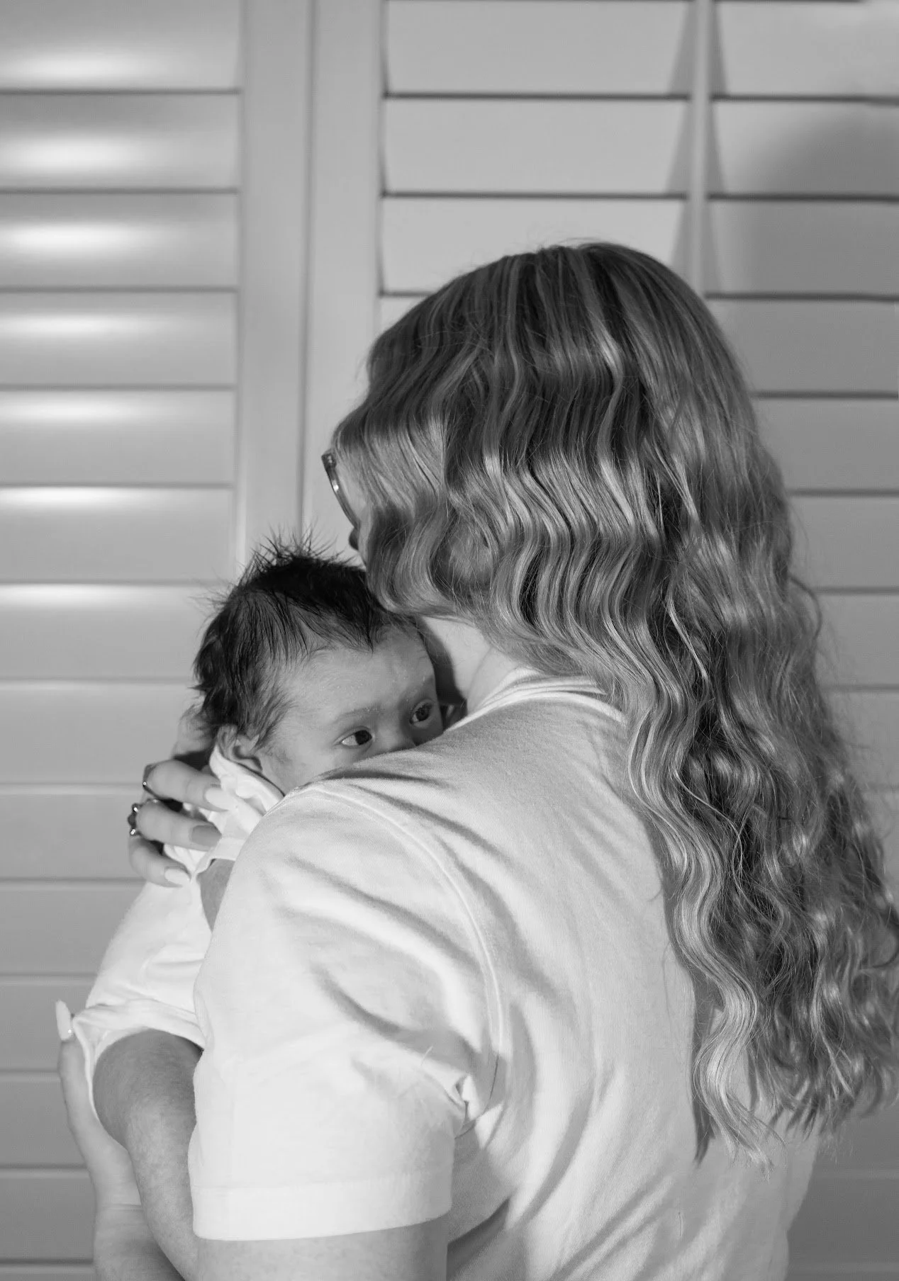 A woman holding a newborn baby close to her face, with her face partly visible, in front of a wall with horizontal panels.
