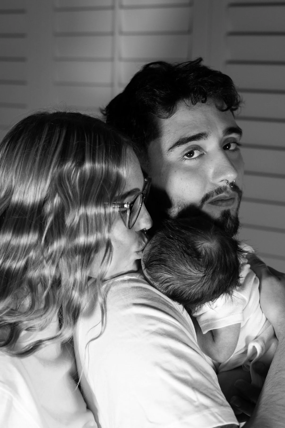 A woman with glasses and long wavy hair is kissing a newborn baby on the head, while the baby is in the arms of a man with a beard and dark hair.