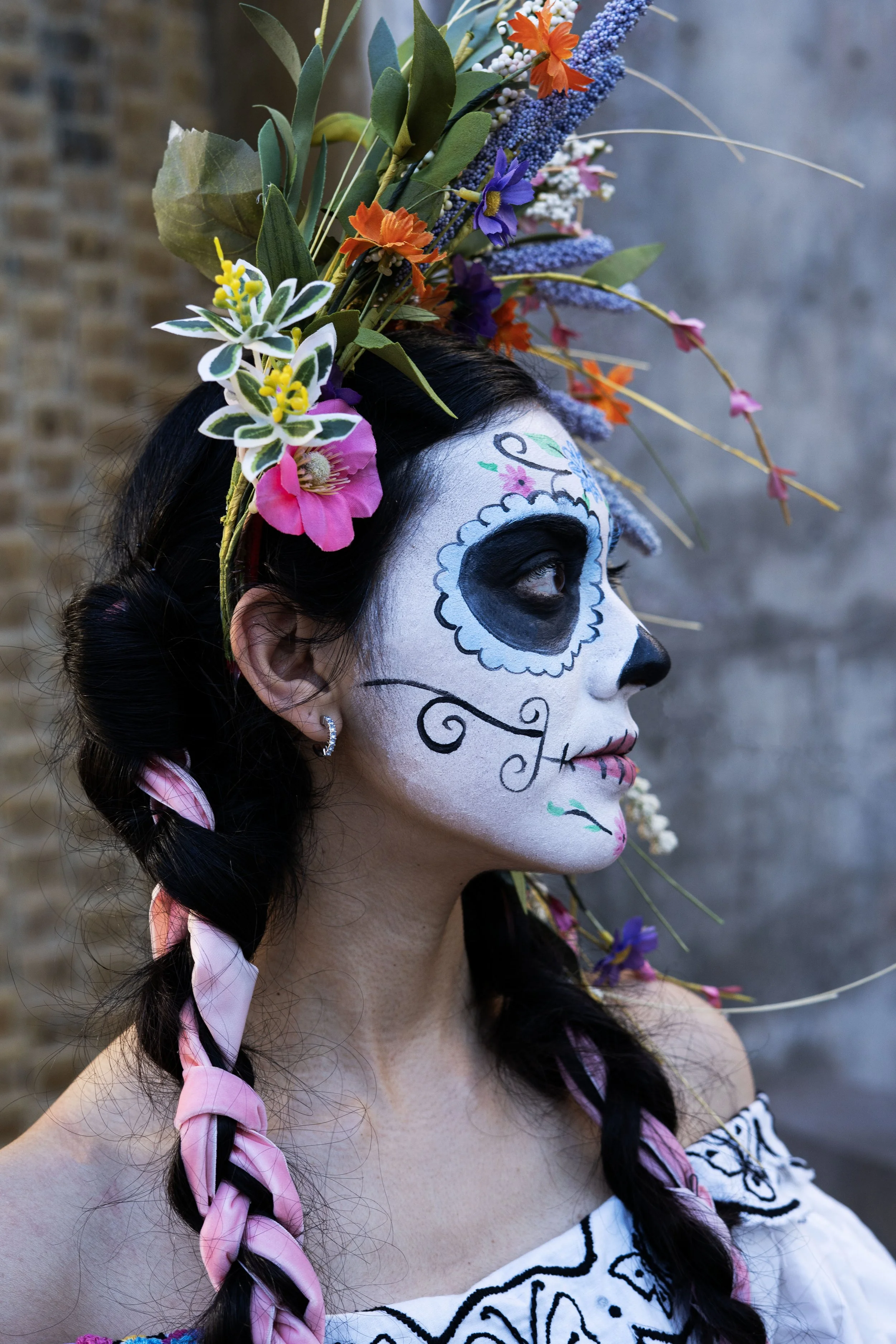 Woman with traditional Day of the Dead face paint, wearing a floral headdress and braid with pink ribbon, dressed in embroidered white blouse, standing against a brick wall.