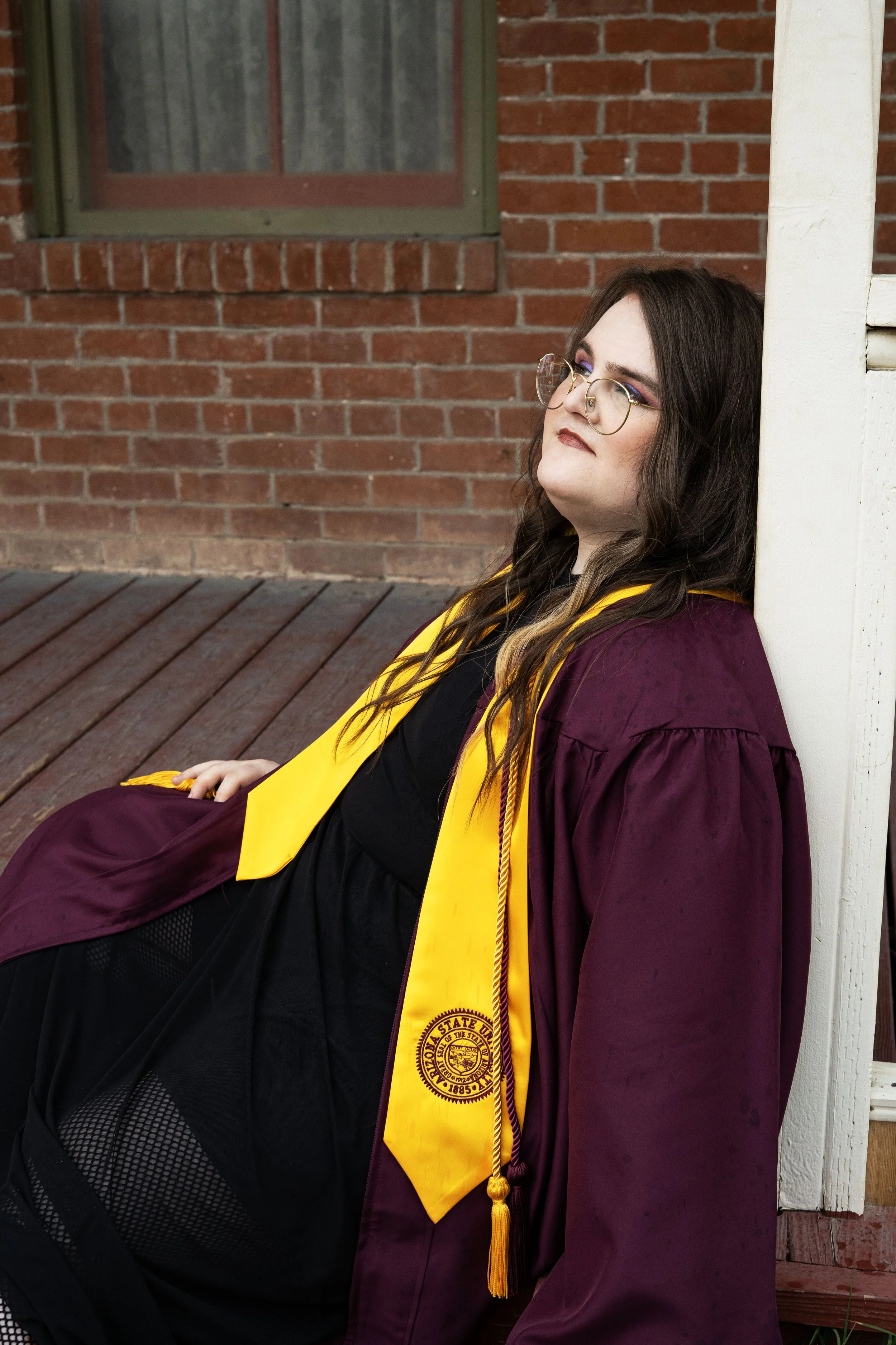 Woman in maroon graduation gown and yellow stole, sitting against a white post, with a brick building in the background.