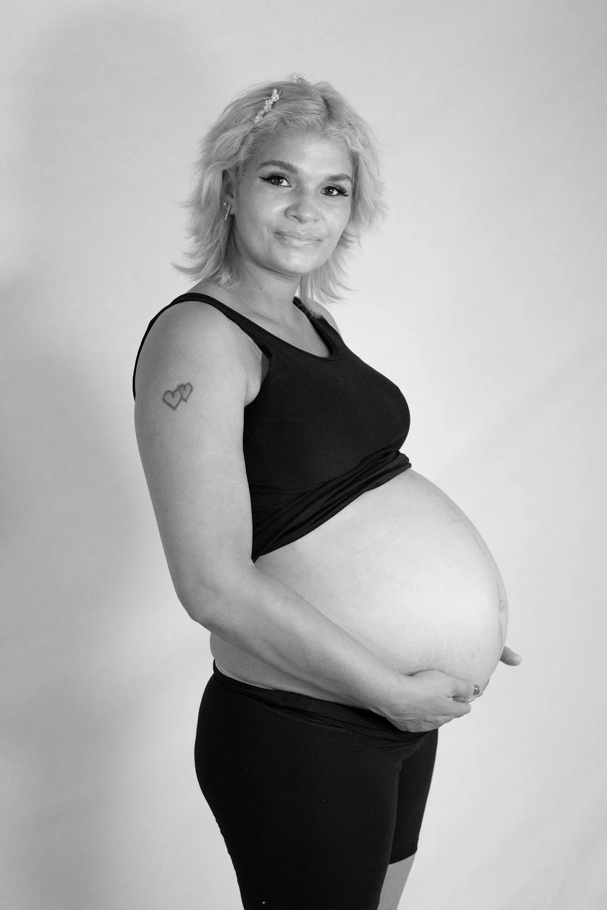 Black and white photo of a pregnant woman with blonde hair, smiling, wearing a black sleeveless top and black pants, standing against a plain background.