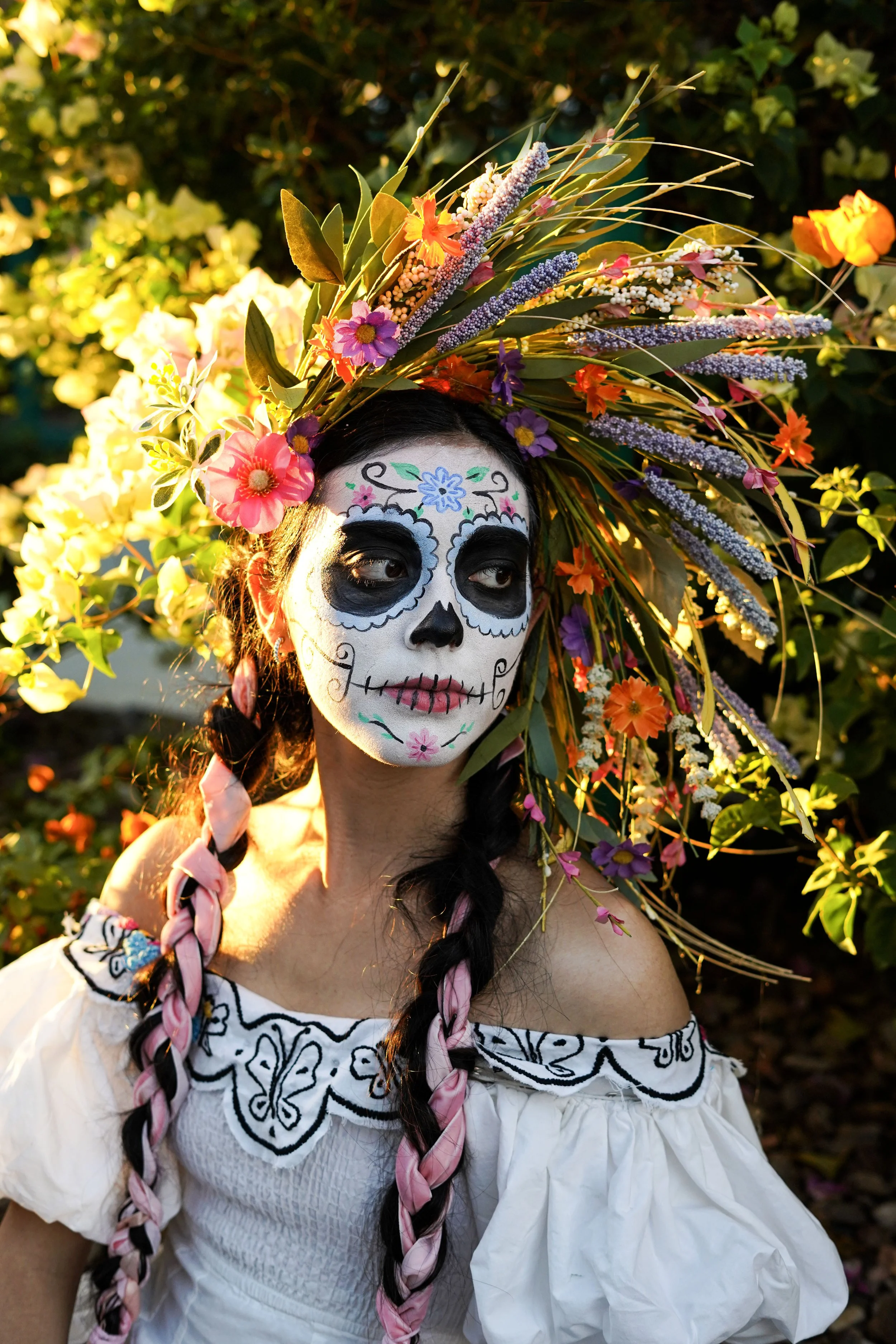 Woman dressed in traditional Mexican attire with sugar skull face paint, wearing a large floral headdress, standing outdoors in sunlight.