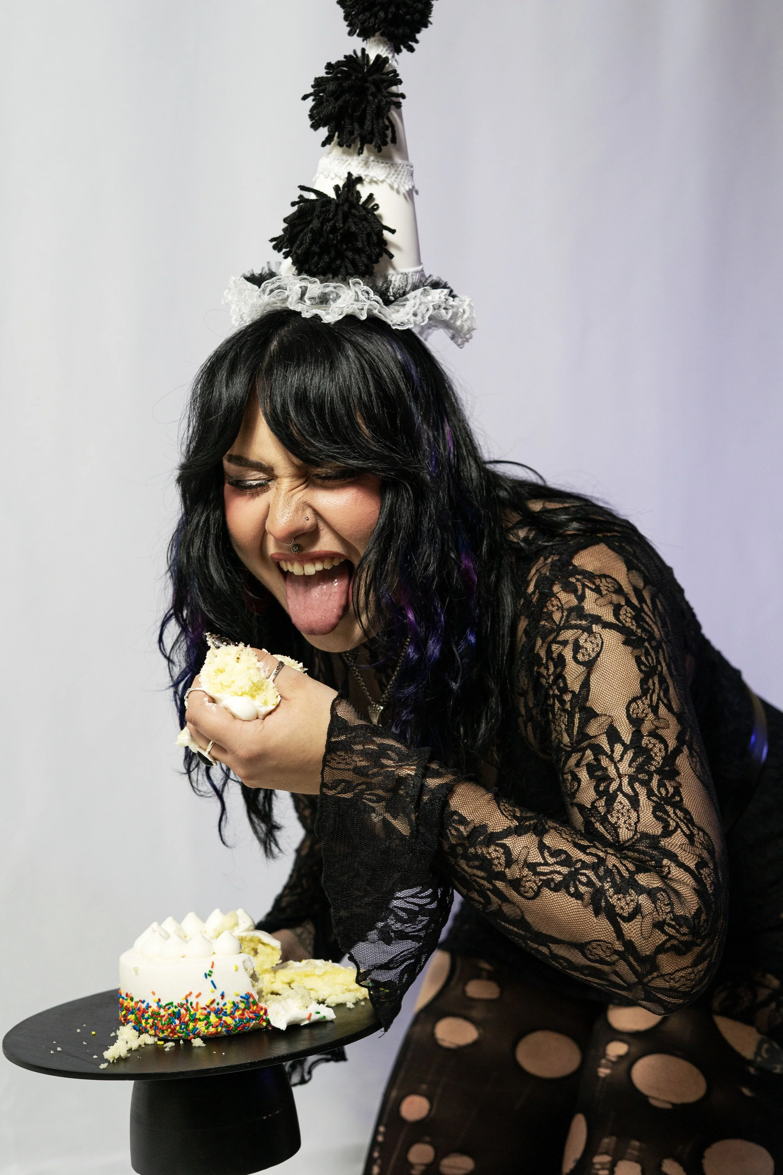 A woman with black hair and purple streaks wearing a lace black dress and a cone-shaped birthday hat decorated with pom-poms and lace, laughing and licking her finger after biting into a slice of cake with rainbow sprinkles, with the rest of the cake