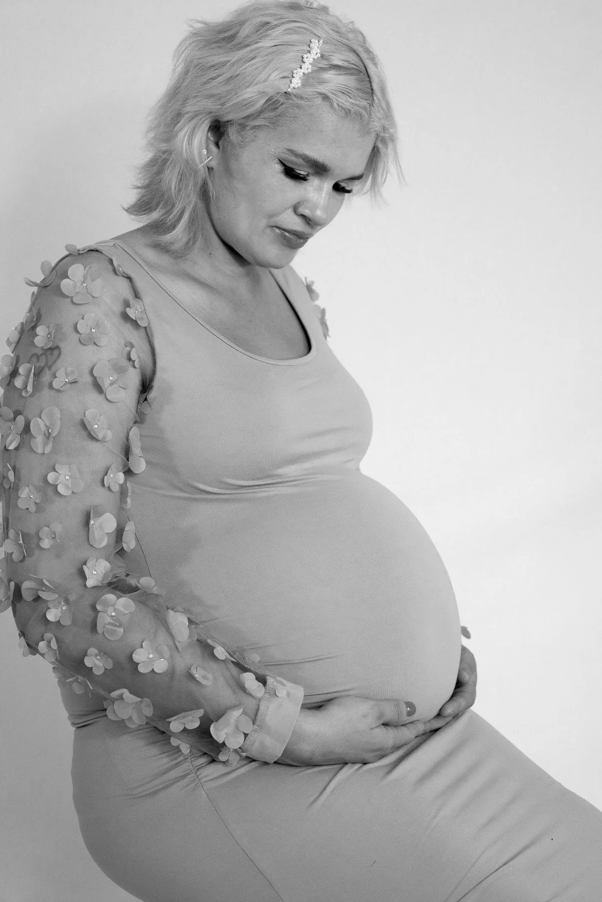 A pregnant woman with short blonde hair and a headband, wearing a dress with floral embellishments on the sleeves, holding her belly and looking down.
