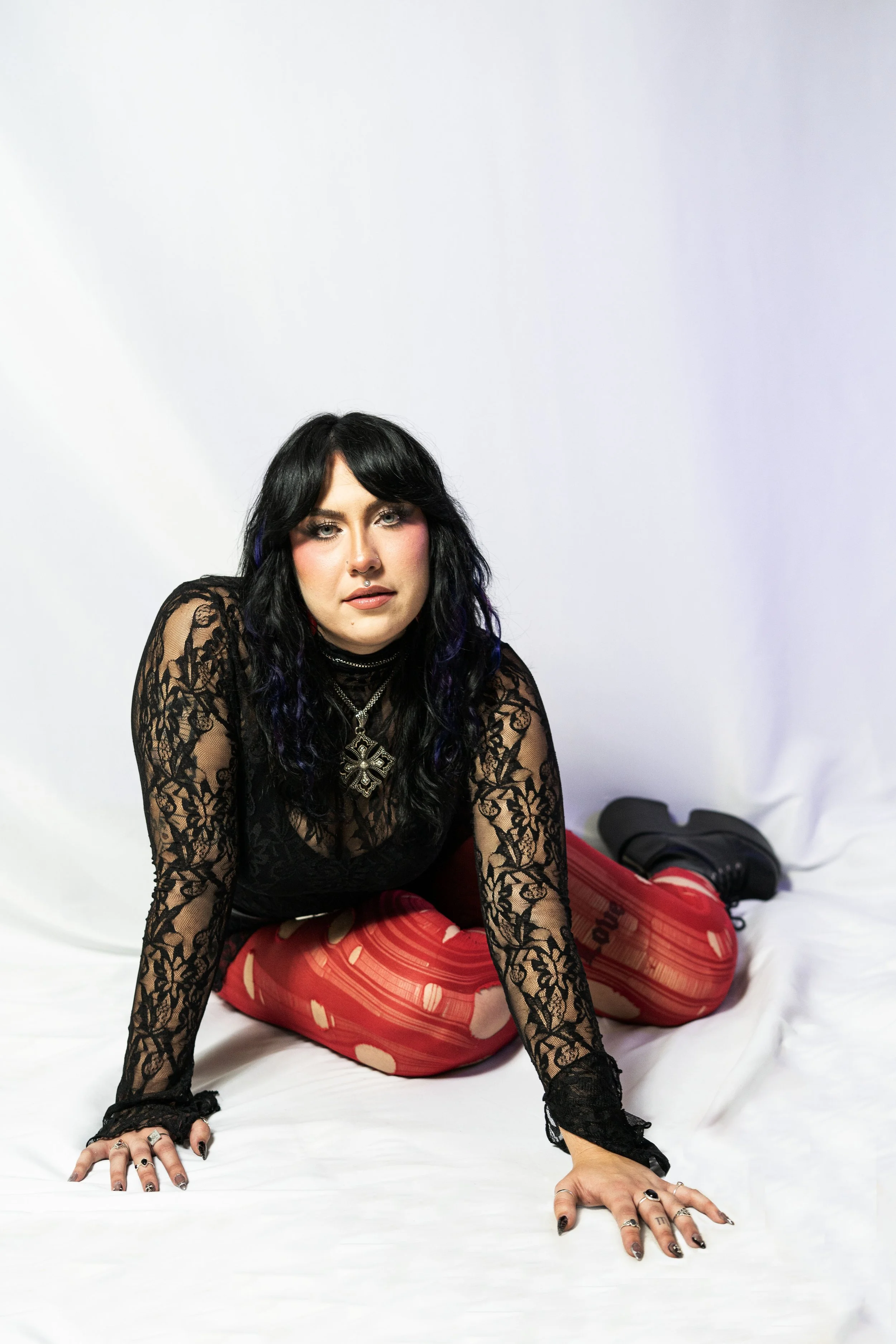 A woman with black hair and purple highlights, wearing a black lace top, red patterned pants, and black boots, sitting on a white surface against a white background.