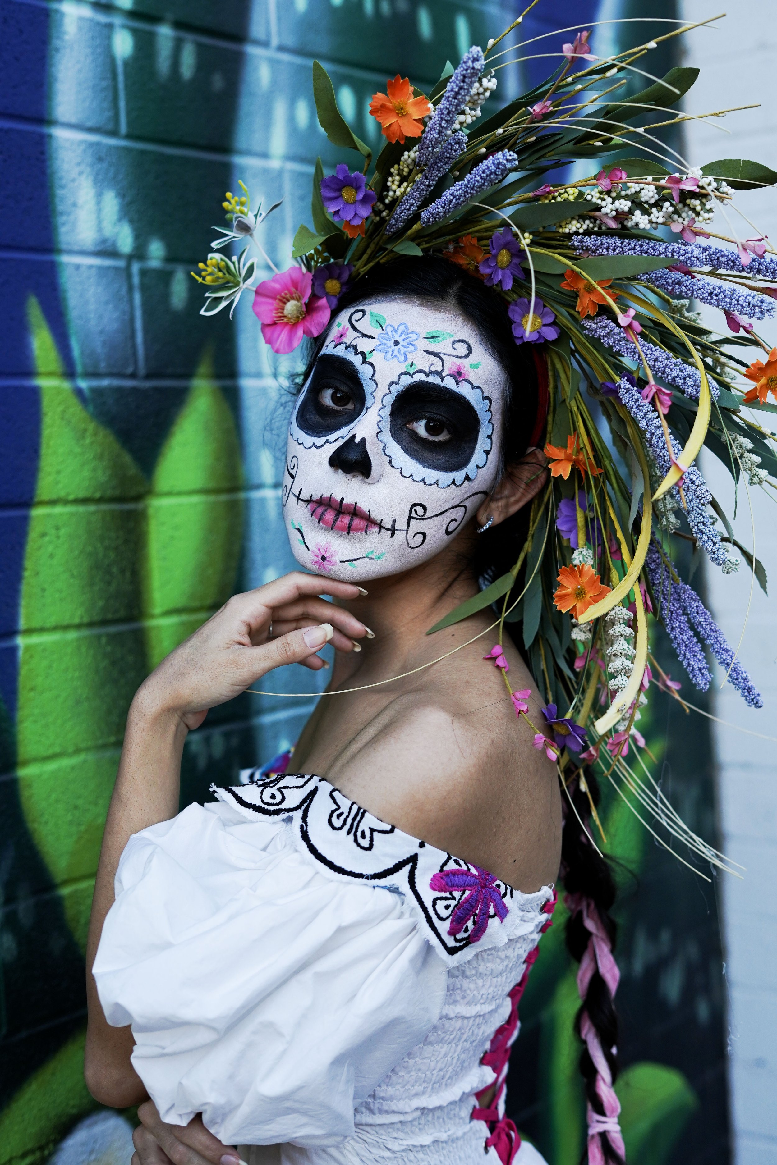 Woman with sugar skull face paint, wearing a floral headdress, standing against a colorful graffiti wall.