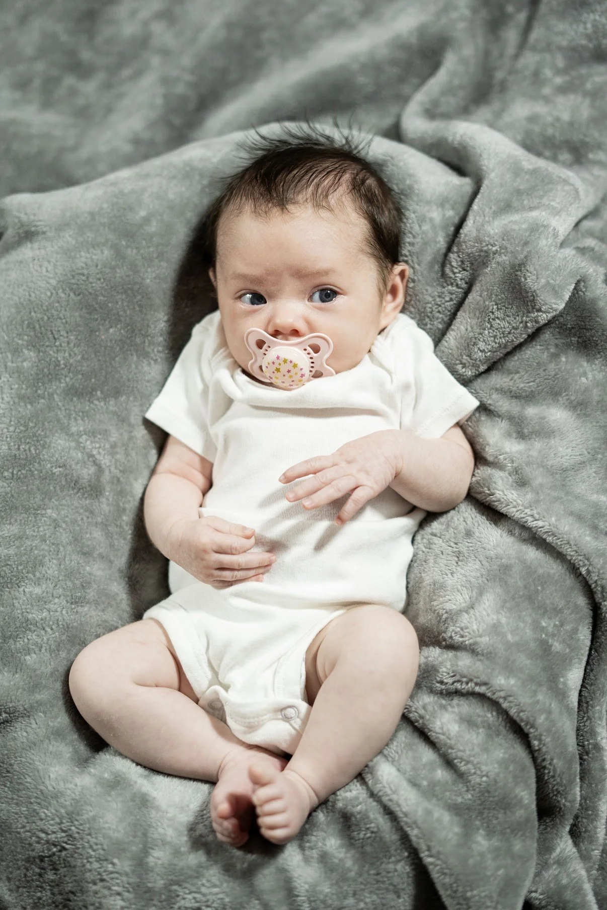 A baby with dark hair and blue eyes lying on a soft gray blanket, wearing a white shirt and a pacifier in mouth.