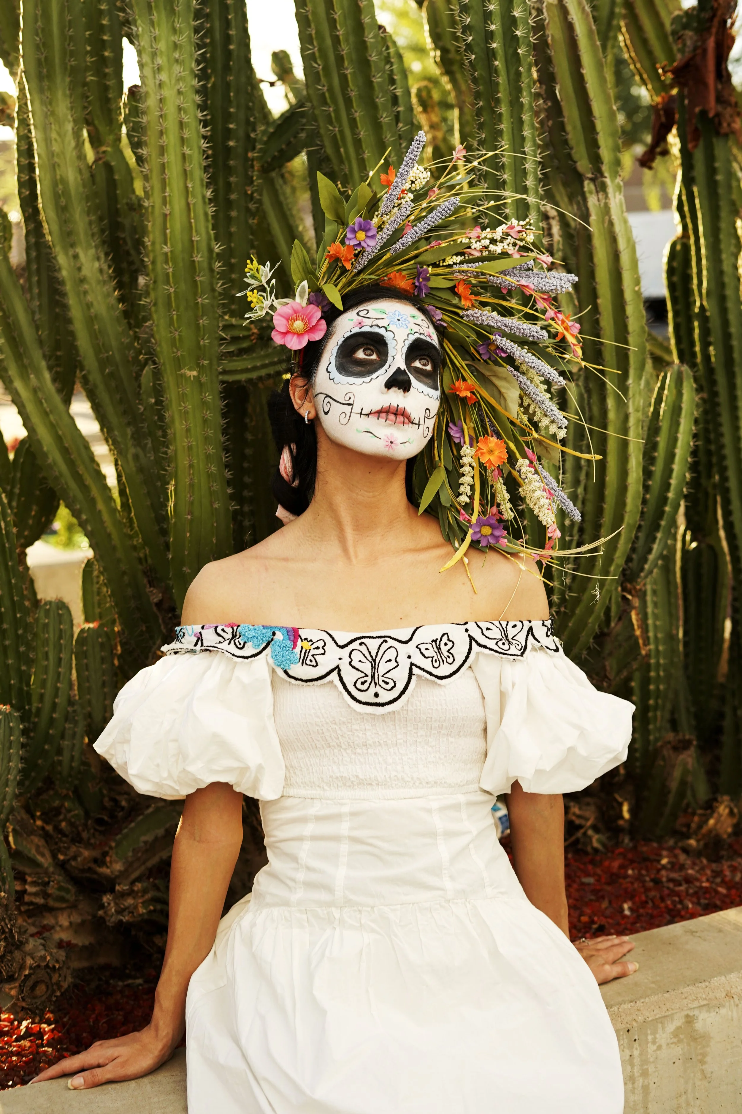 Woman with sugar skull face paint, wearing a white embroidered off-shoulder dress, sitting in front of tall cacti, with a colorful floral headdress, celebrating Day of the Dead.