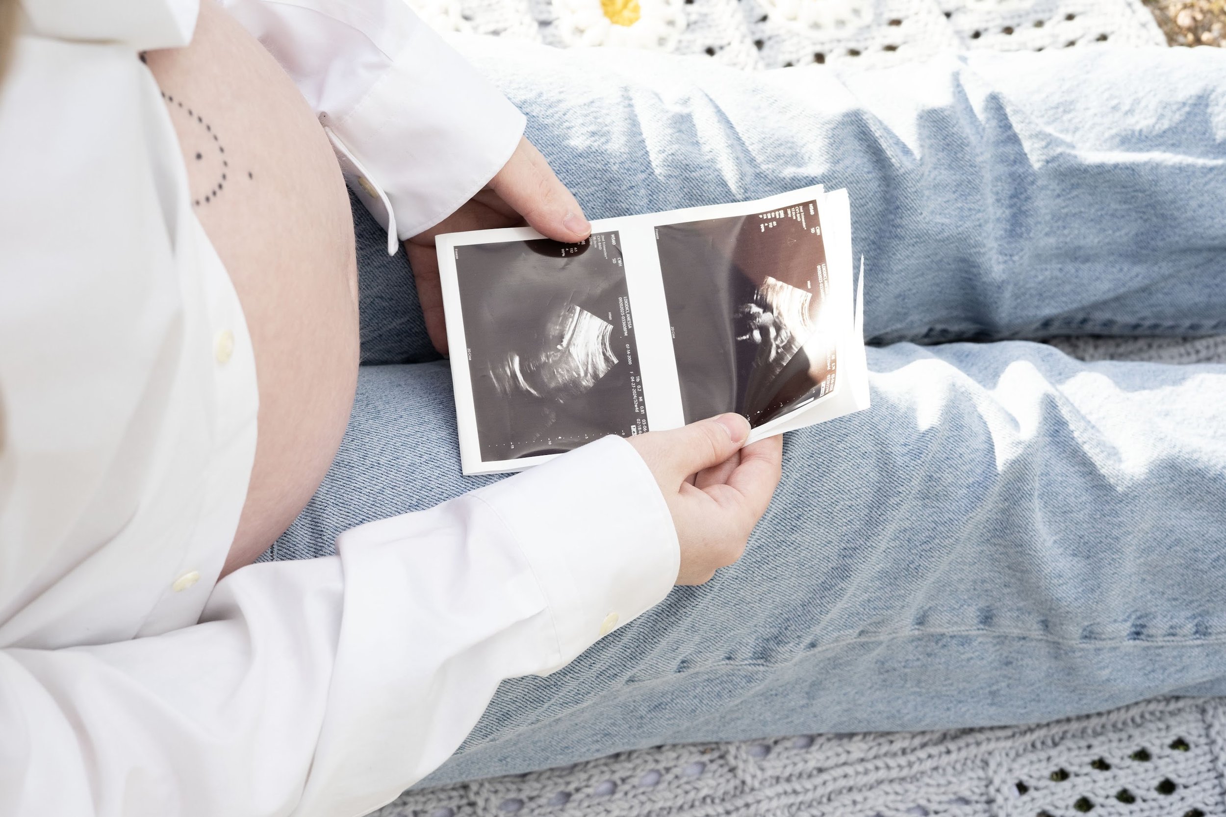 A person holding two ultrasound images while sitting outdoors on a blanket or rug.
