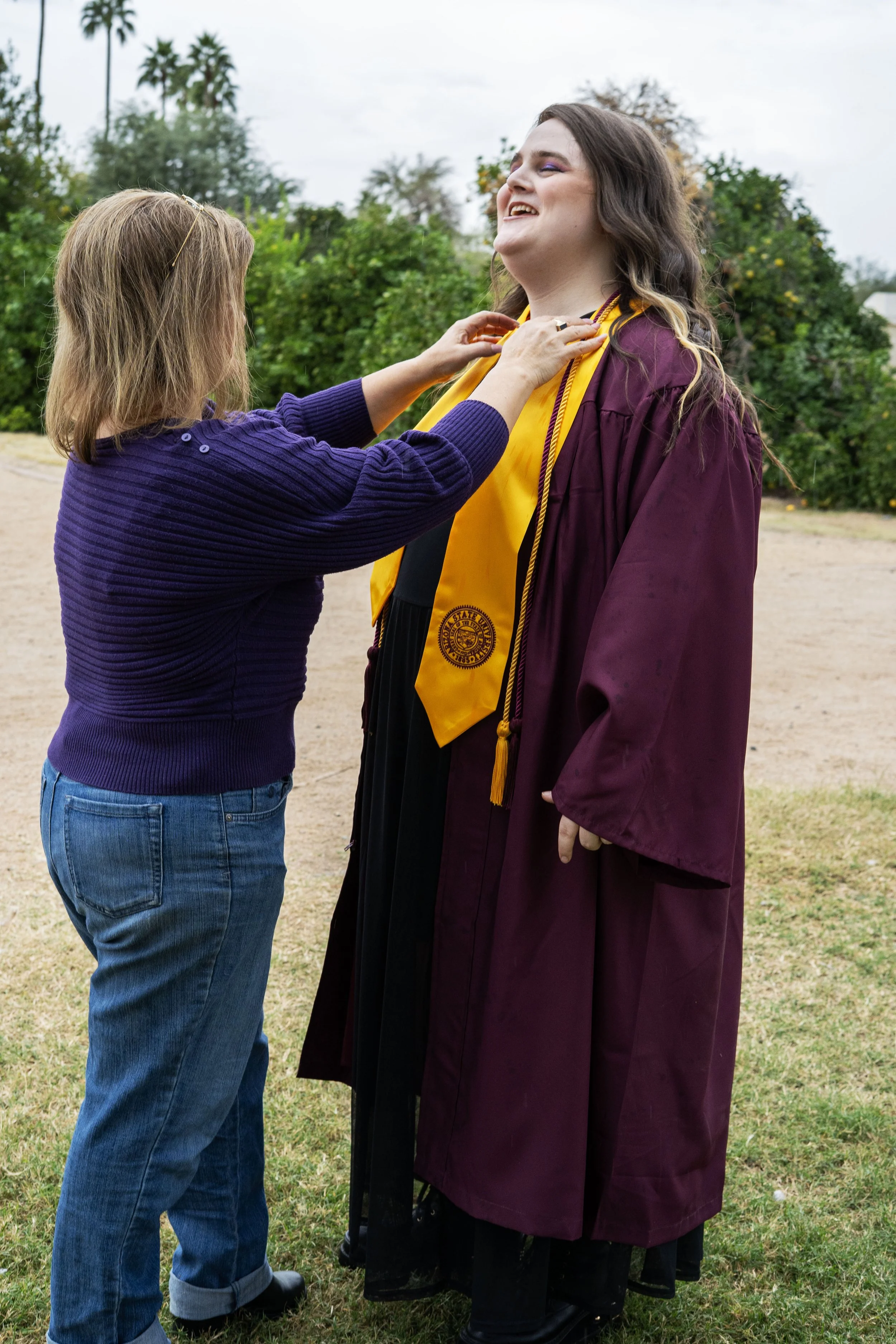 A woman in purple sweater and blue jeans placing a graduation stole on a smiling graduate wearing a maroon cap and gown outdoors.