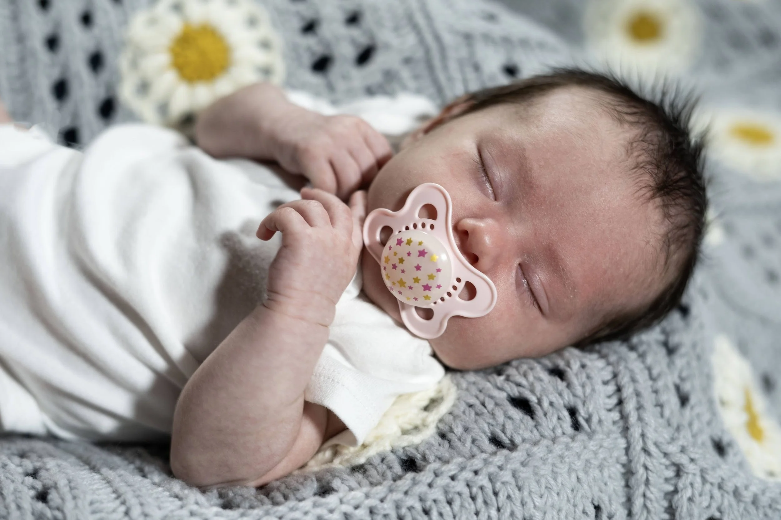 A sleeping baby with a light pink pacifier lying on a gray knitted blanket with white and yellow flowers.